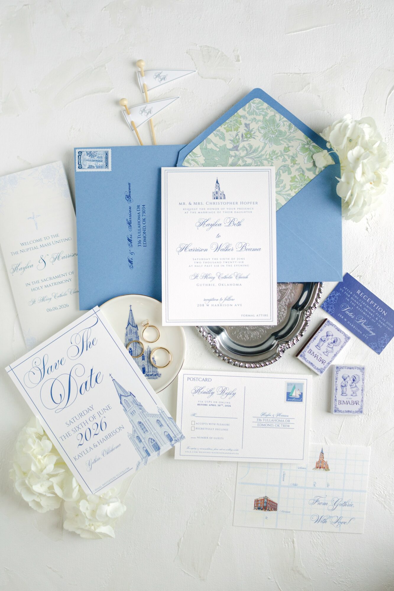 Wedding invitation, blue envelope, white flowers, jewelry, and stationery on a white surface.