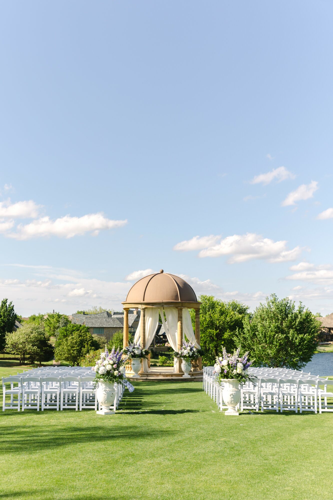 Outdoor wedding setup with a gazebo, chairs, and floral arrangements on a grassy field under a blue sky.