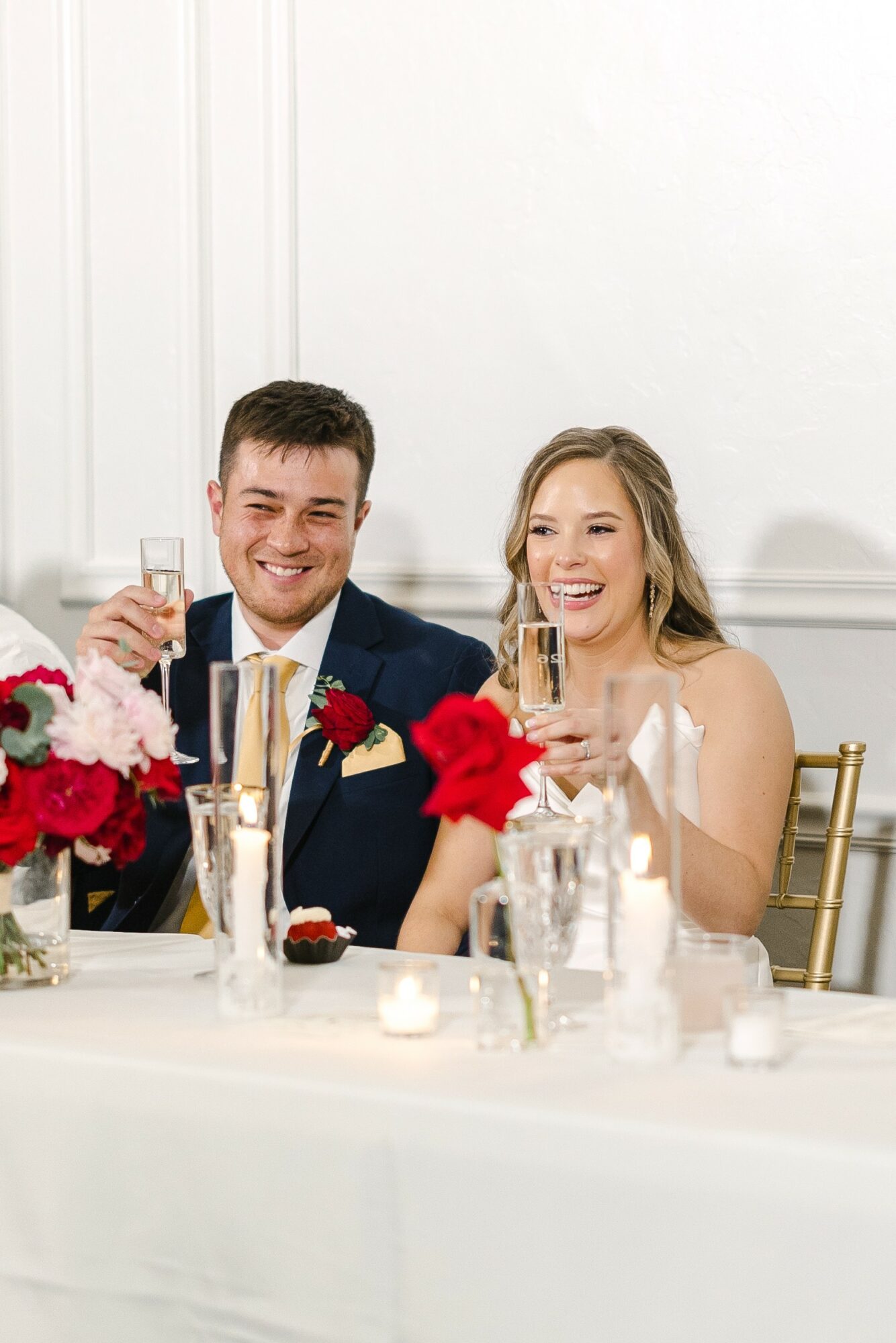 A smiling couple at a wedding reception, holding champagne glasses, sitting at a decorated table with flowers and candles.