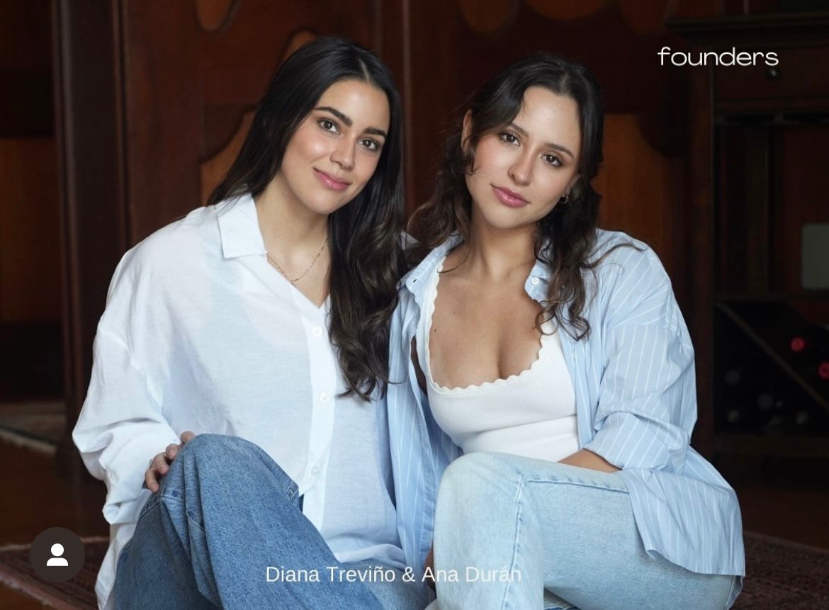 Two women sit close together, smiling, in a wooden-paneled room, wearing casual white and blue clothing.
