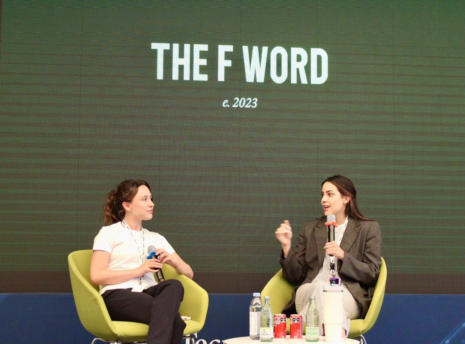 Two women sit on stage in yellow chairs, engaging in conversation with a large screen behind them displaying 'THE F WORD' and 'c. 2023'.