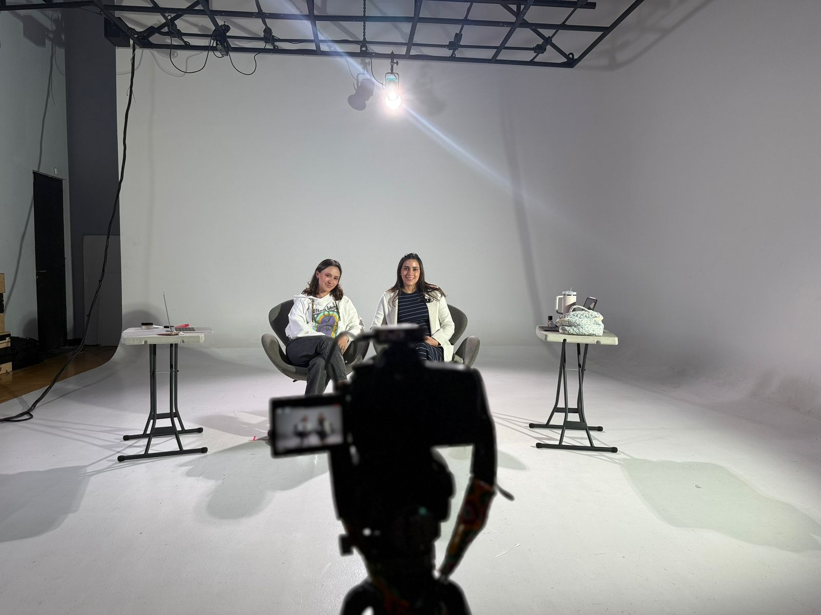 Two women sit at tables in a white studio with a camera in foreground, bright light overhead, and minimal background.
