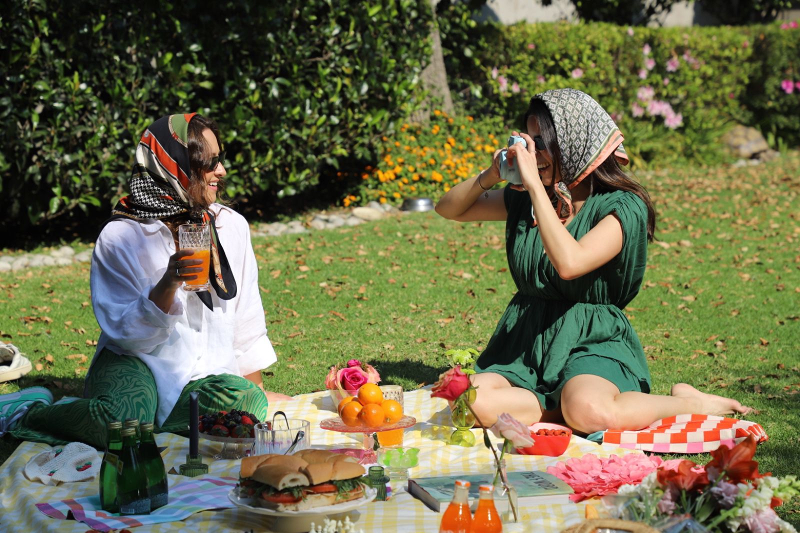 Two women sitting on a picnic blanket in a garden, surrounded by food and flowers, one holding a camera and the other with a drink.