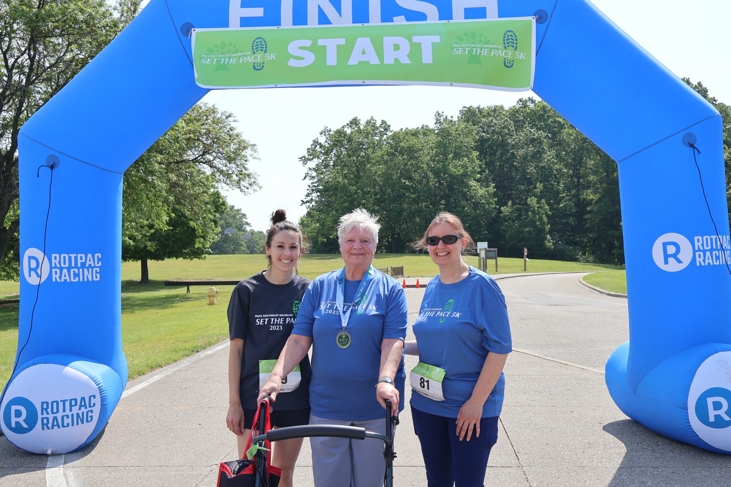 Three women standing under a blue inflatable start arch at a race event, smiling, with trees in the background.