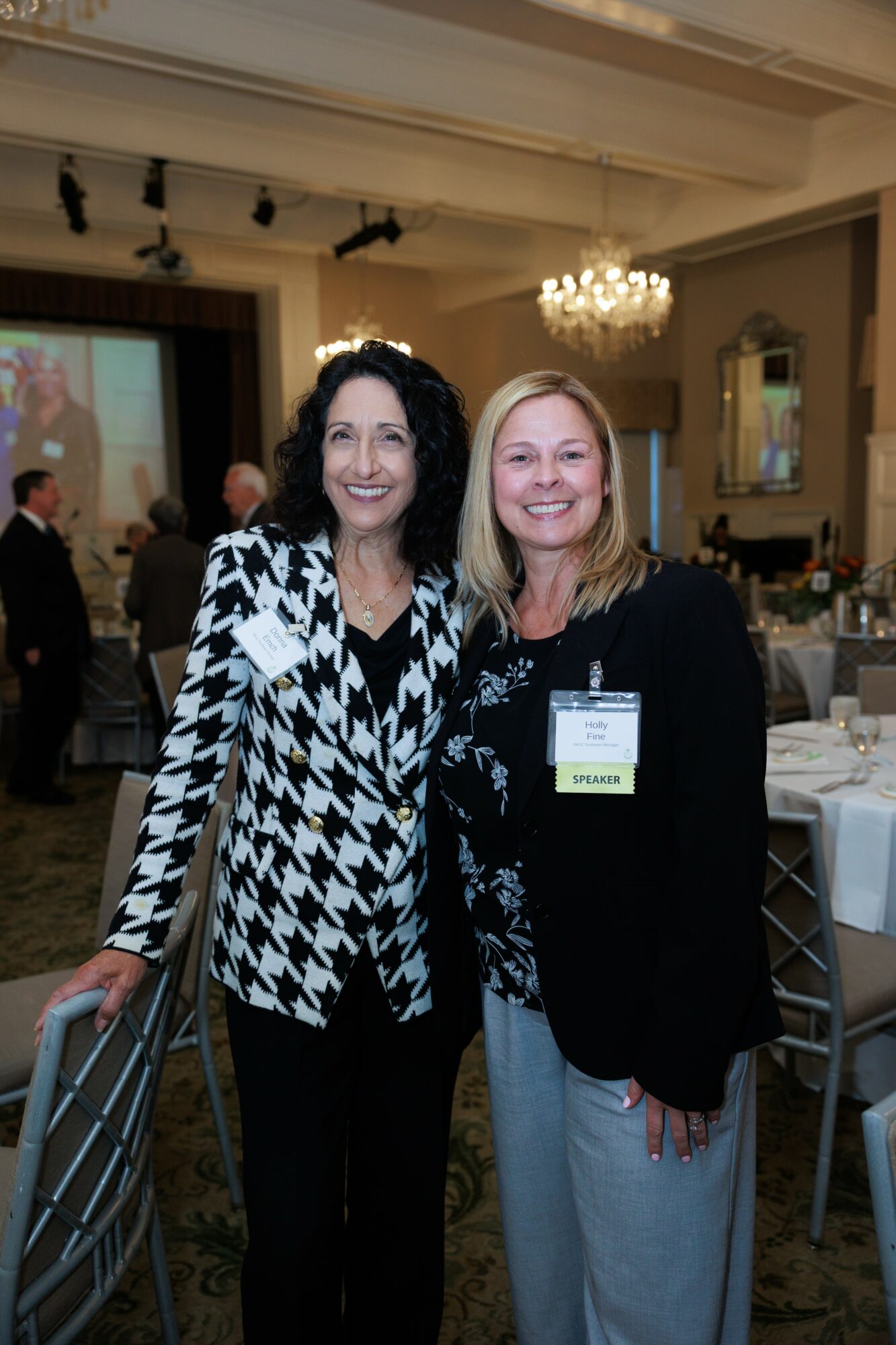 Two women smiling and standing together in a banquet hall with tables and a chandelier.