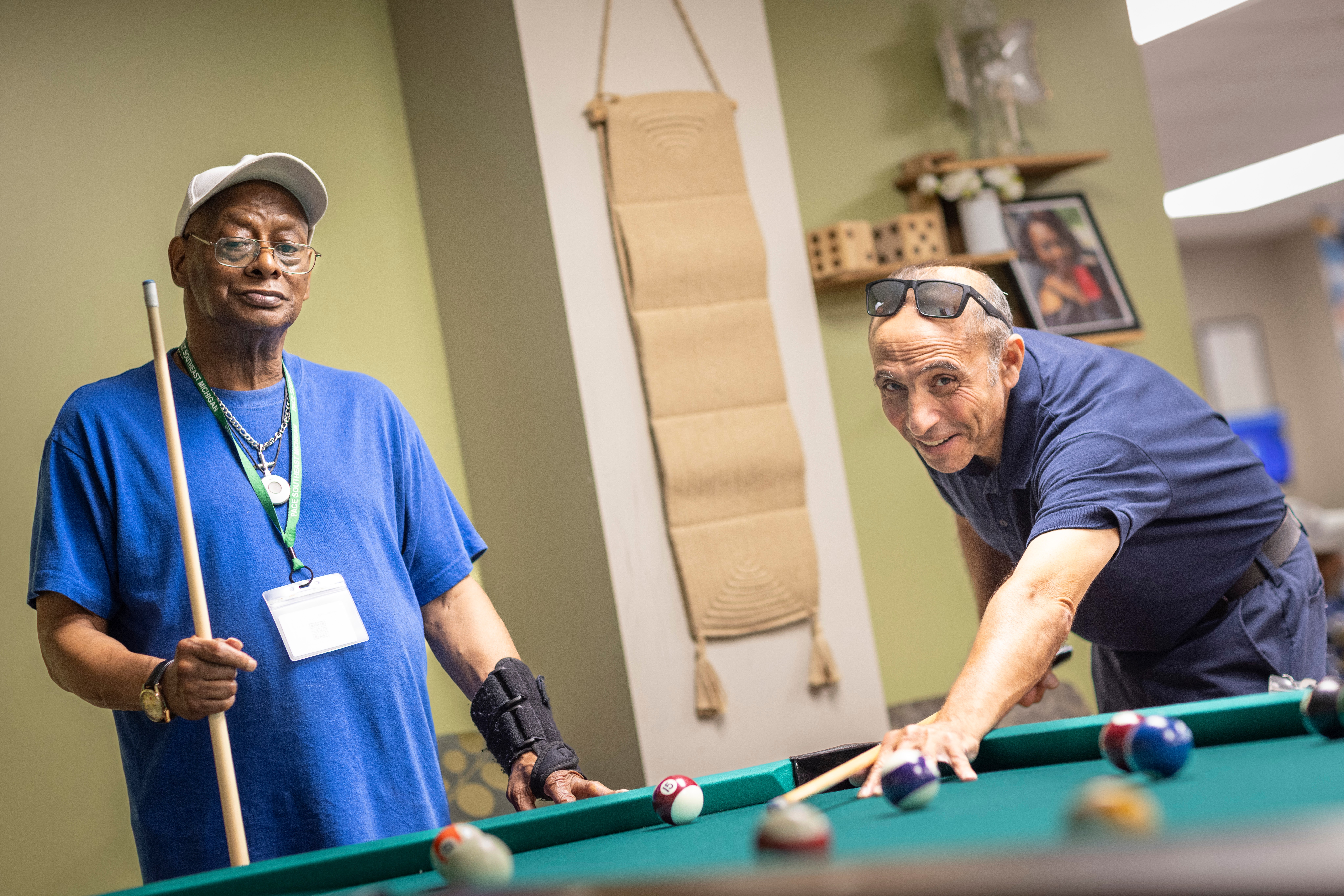 Two elderly men play pool indoors, one wearing a blue shirt and glasses, the other in a dark shirt, leaning over the table.