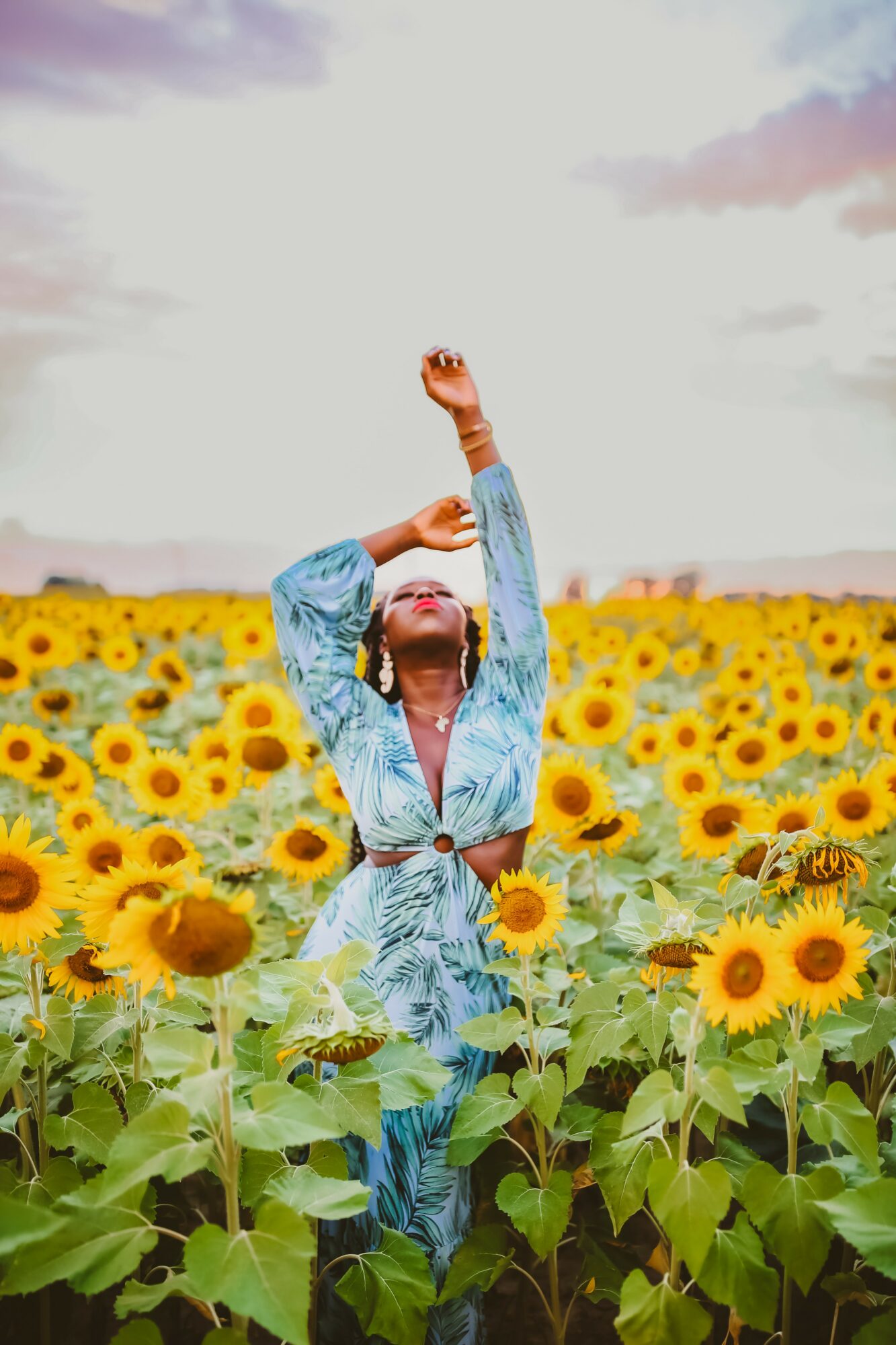 Person standing in a sunflower field with arms raised, wearing a blue outfit, under a partly cloudy sky.