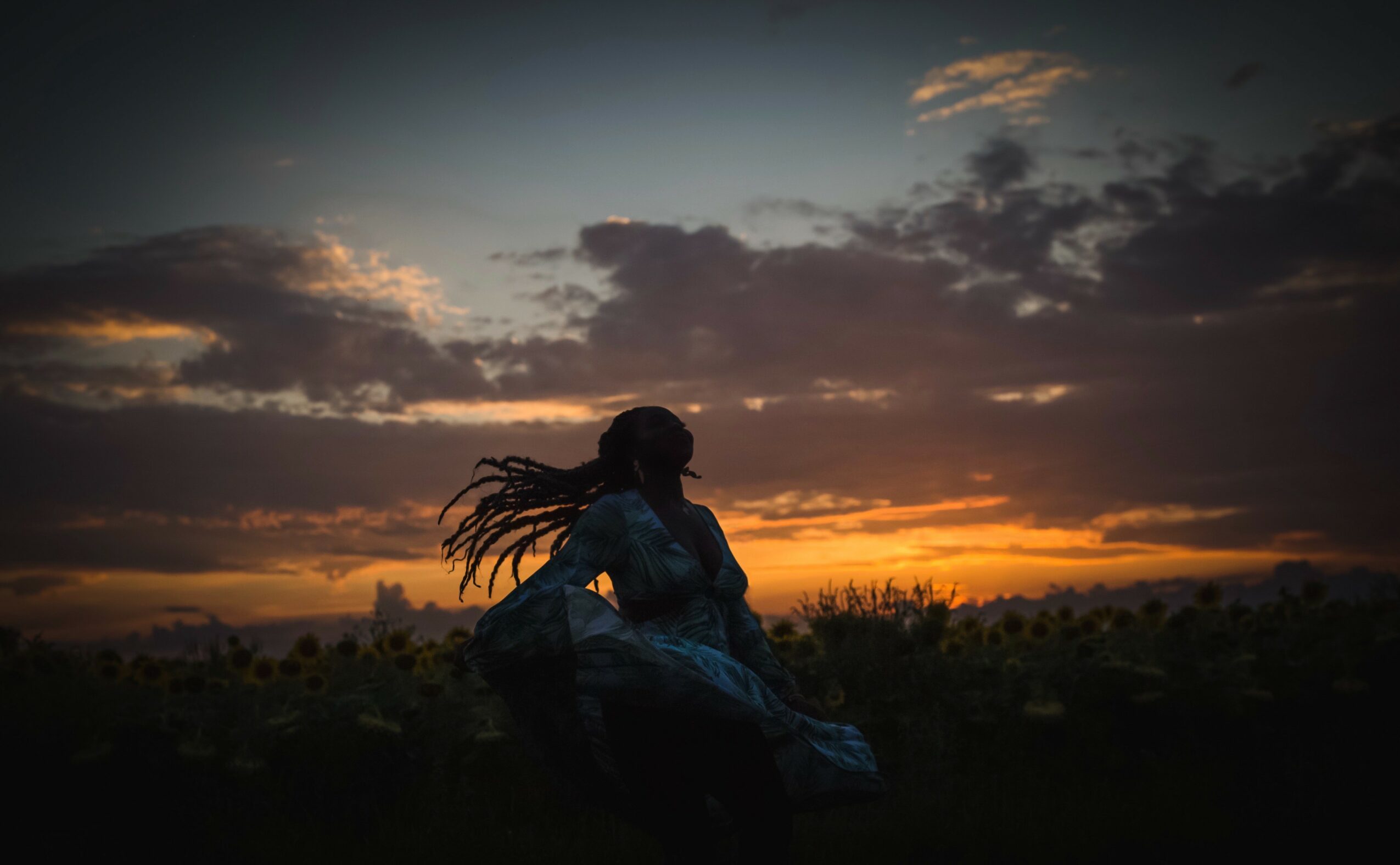 Silhouette of a person with flowing hair sitting outdoors during sunset with a colorful sky and clouds.