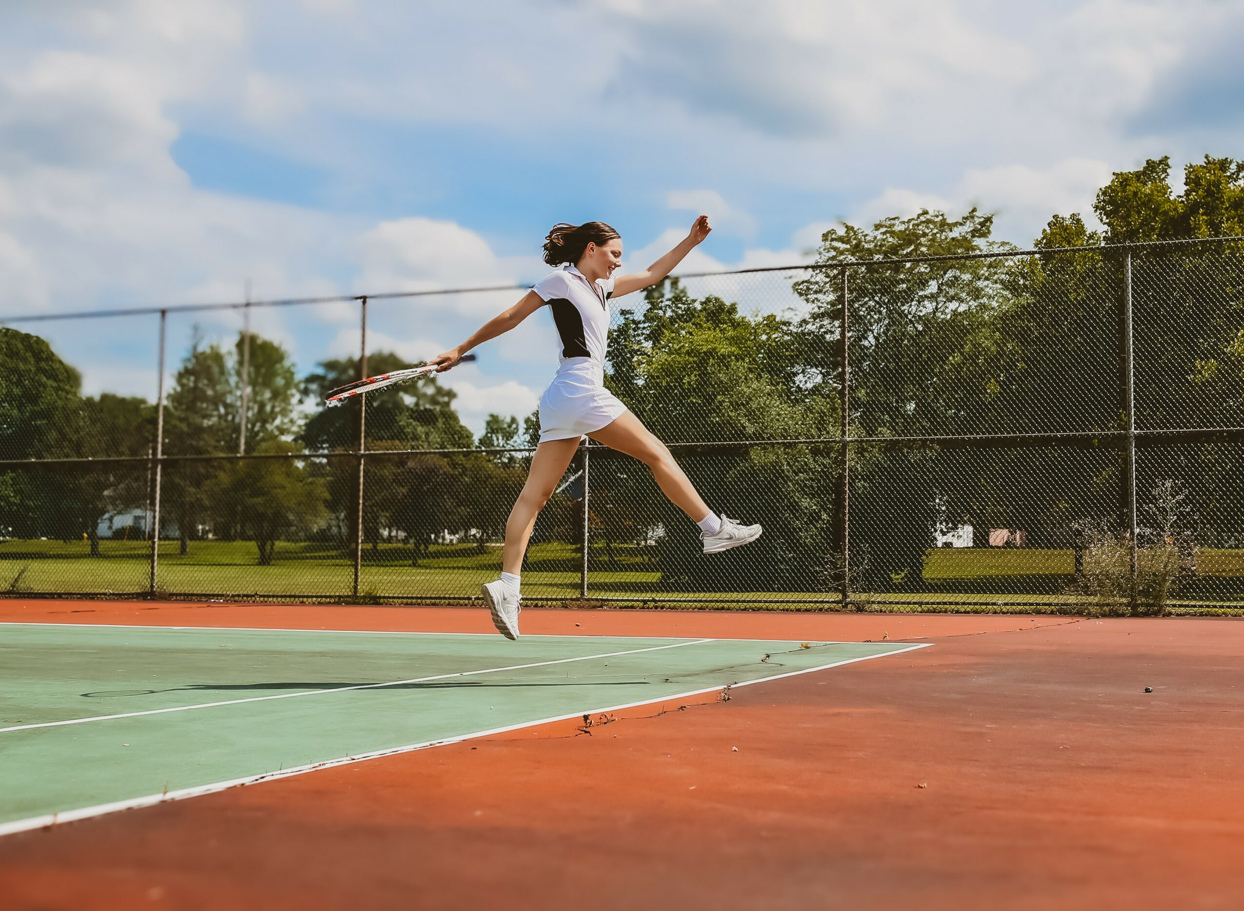Young woman jumping over a tennis net on an outdoor court, with trees and a fence in the background.