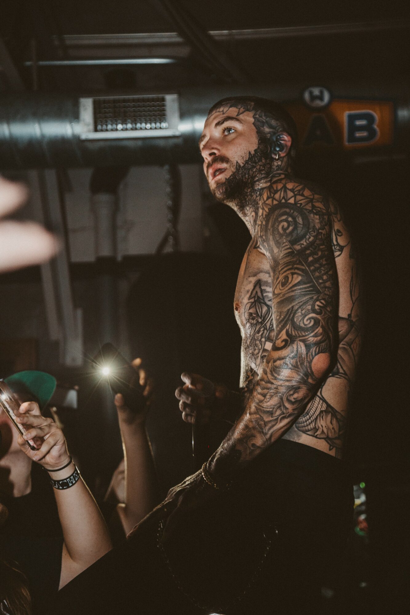 Tattooed man with a beard on stage, looking upward, surrounded by audience, in a dark indoor setting.