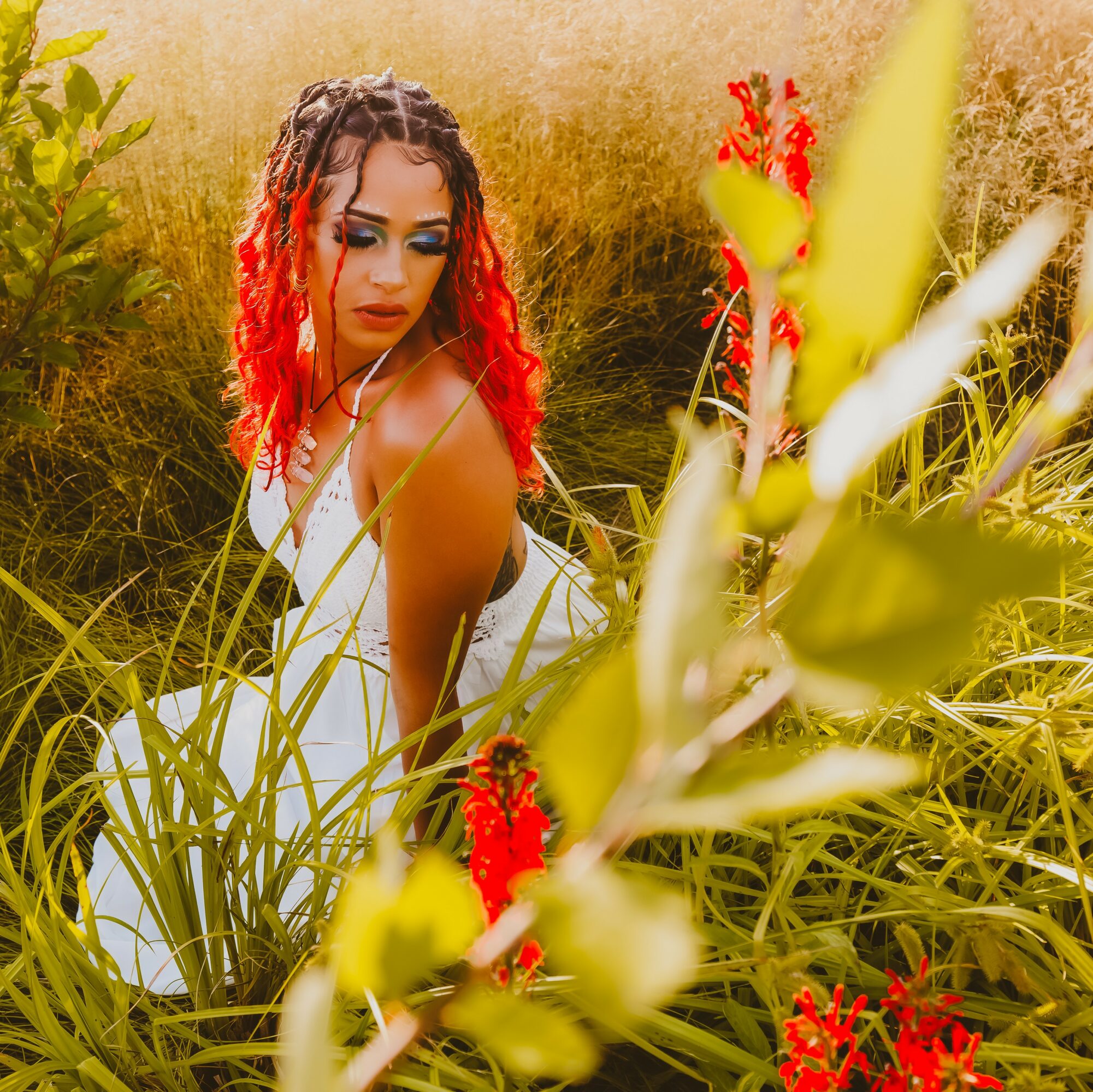 Woman with curly hair and red highlights among tall green plants and red flowers outdoors.