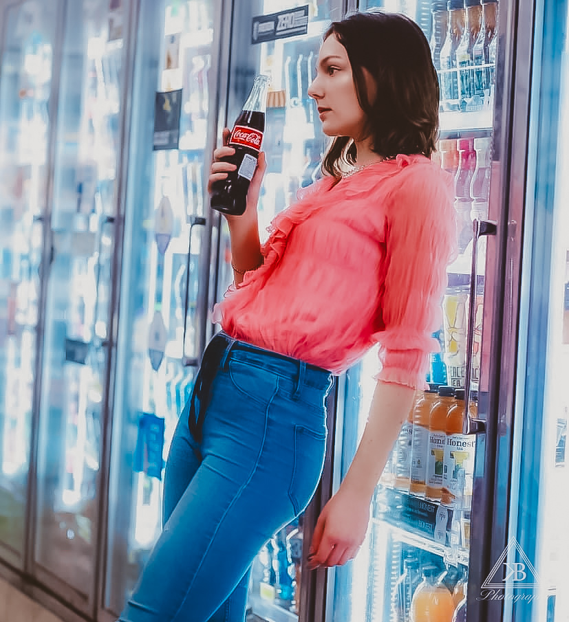 Woman in pink blouse and blue jeans holding a soda bottle in front of vending machines, looking to the side.