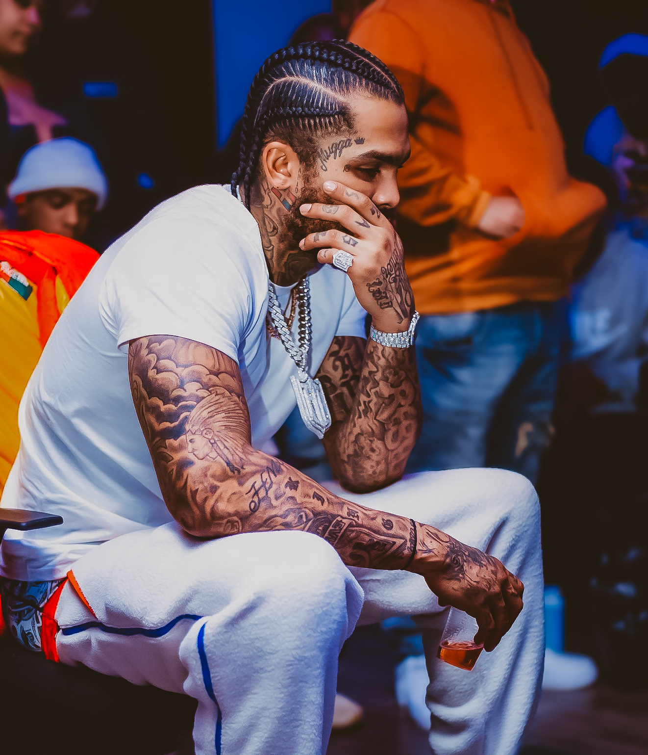 Person with braided hair and tattoos, sitting with hand on face, wearing white clothing, in a dimly lit environment.