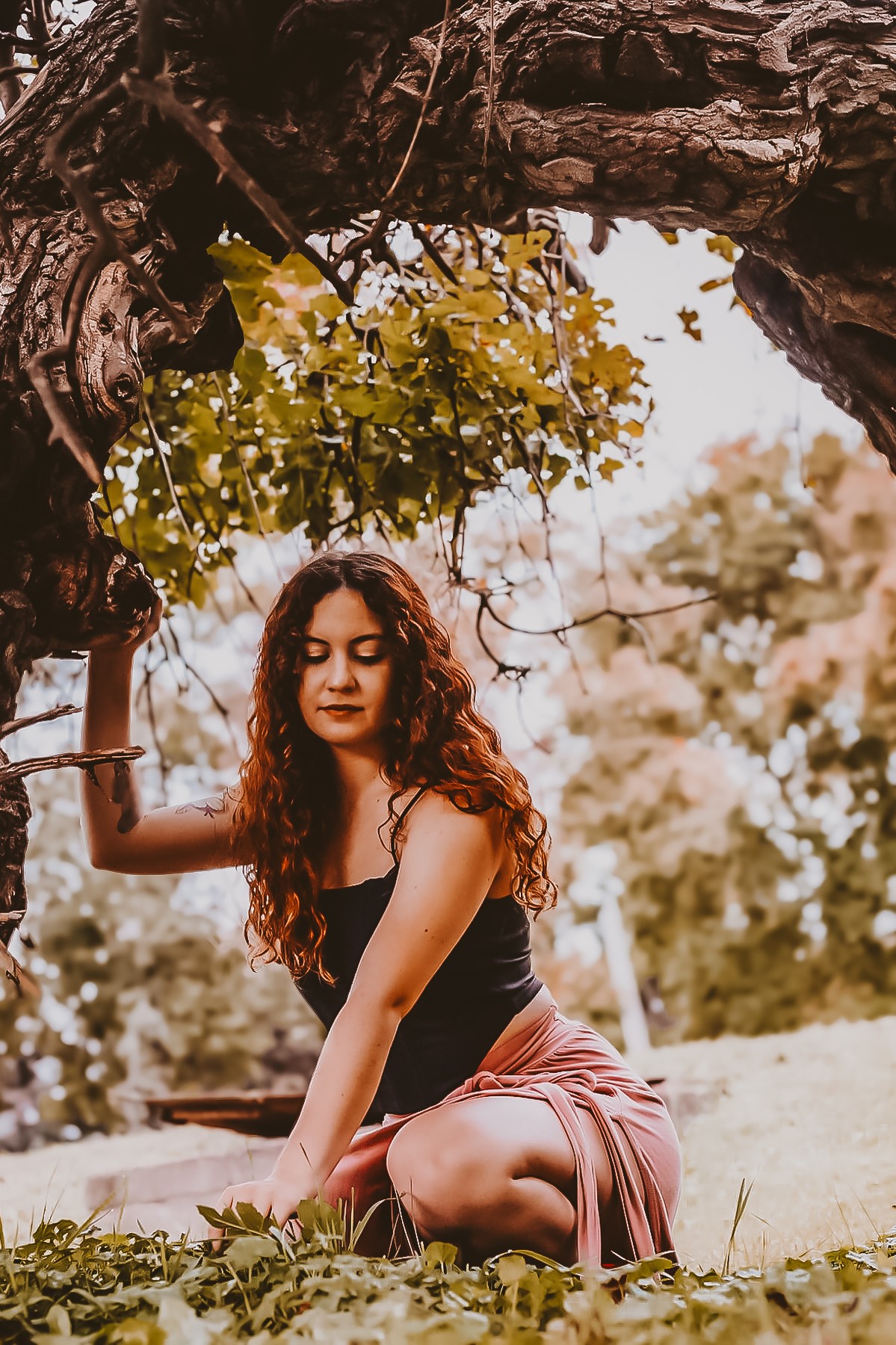 Young woman with curly hair sitting outdoors under a tree, surrounded by greenery and sunlight, looking down thoughtfully.