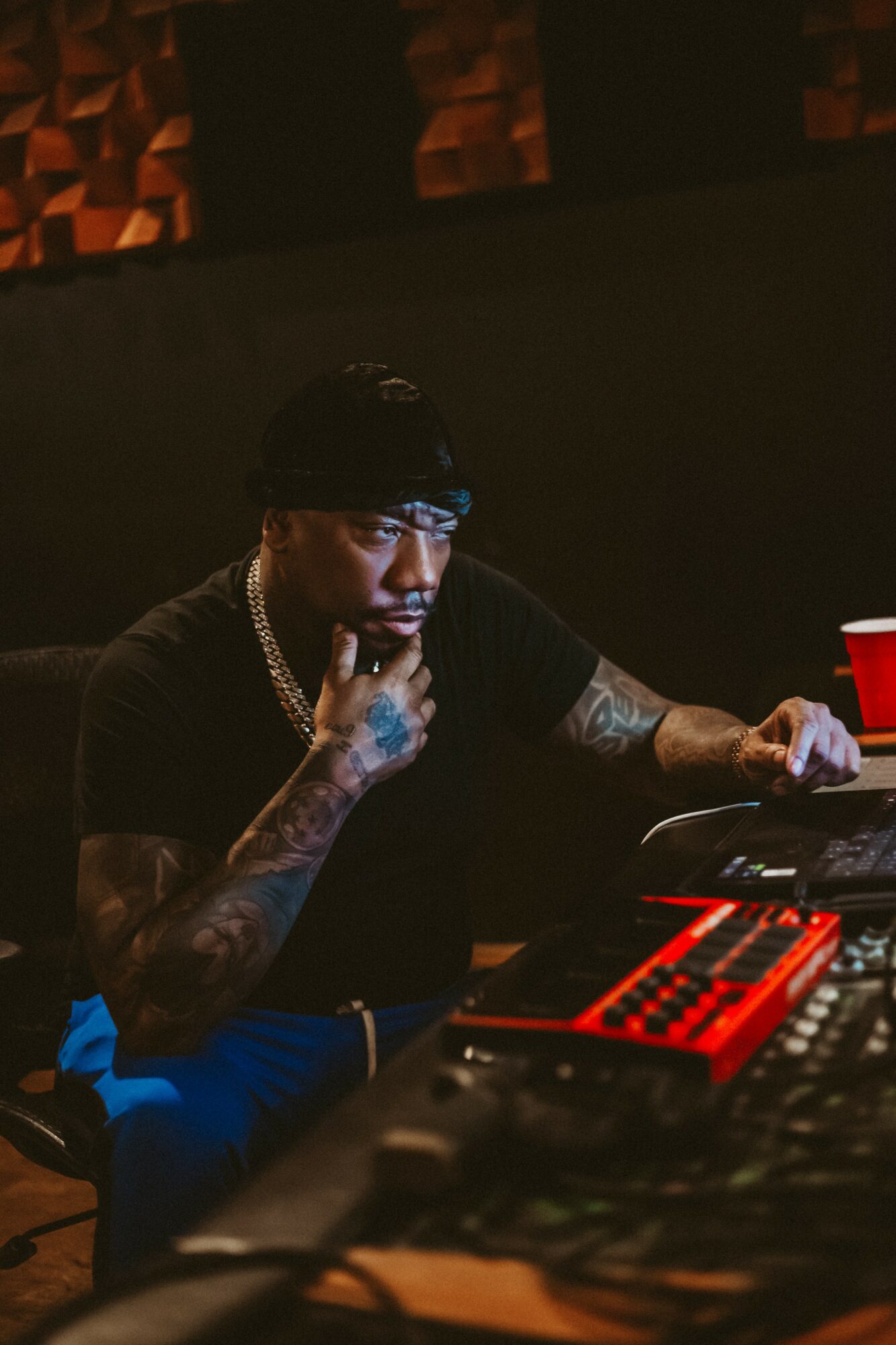 Man with tattoos, wearing a cap and chain, working on a music keyboard in a dark room with red lighting.