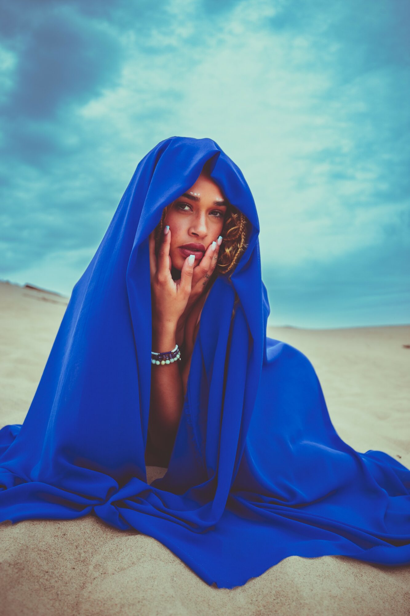 Woman sitting on sand, covered with a blue fabric, under a cloudy sky, touching her face with her hand.