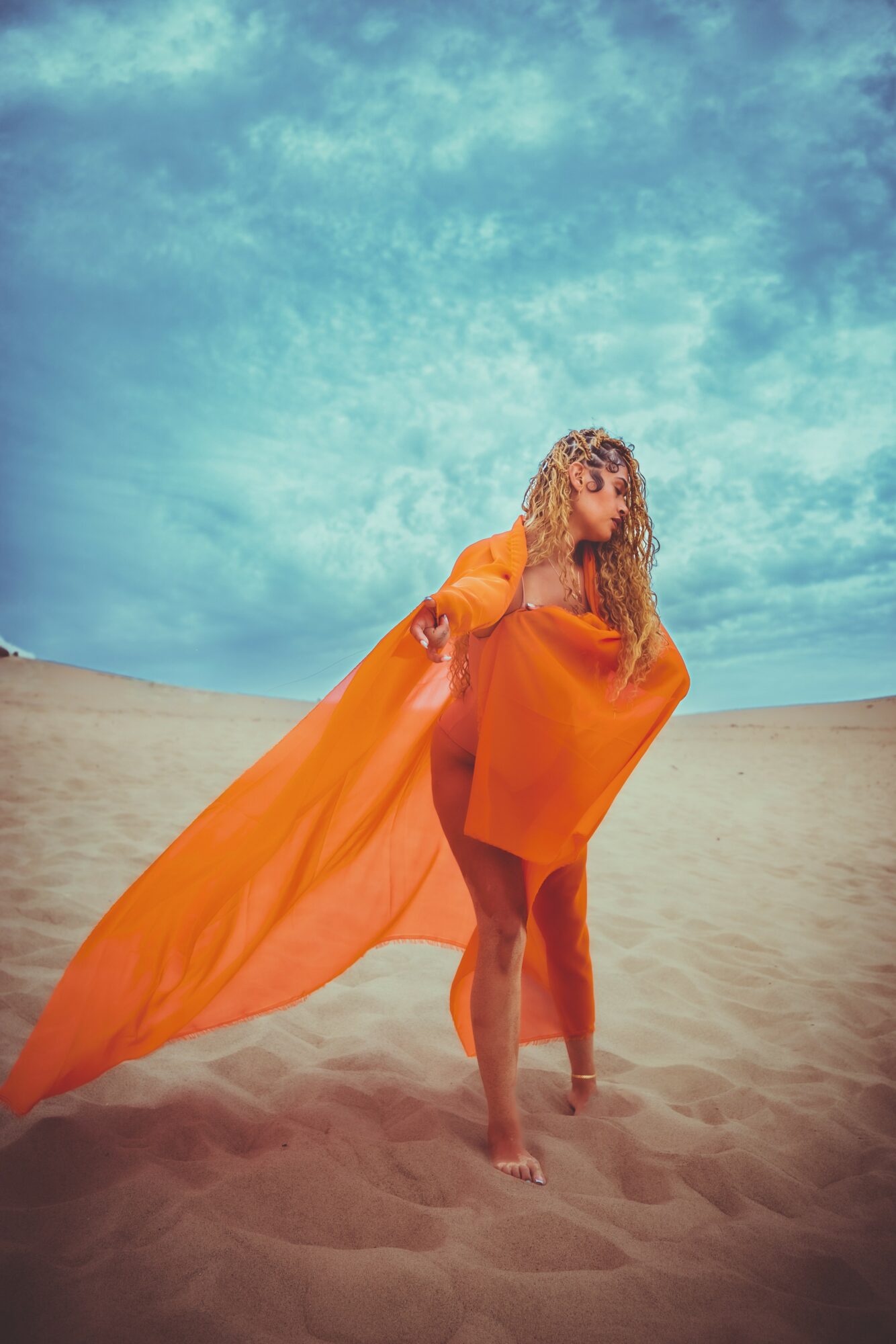 Woman in orange dress standing on sandy desert with blue sky background, holding flowing fabric behind her.