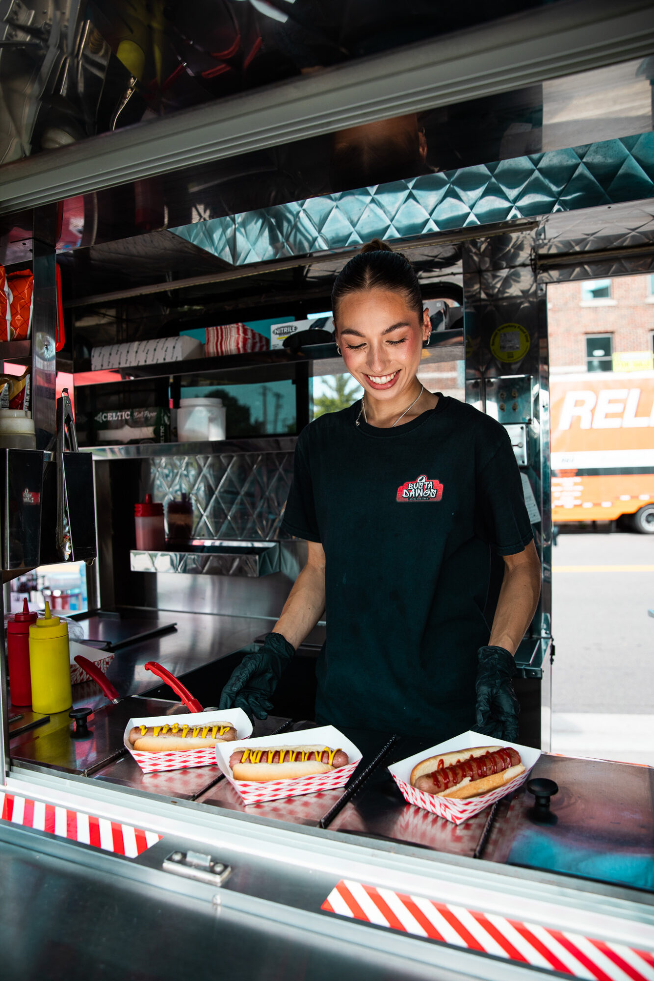 Smiling woman working at a food stand with hot dogs and condiments, inside a food truck or stall.