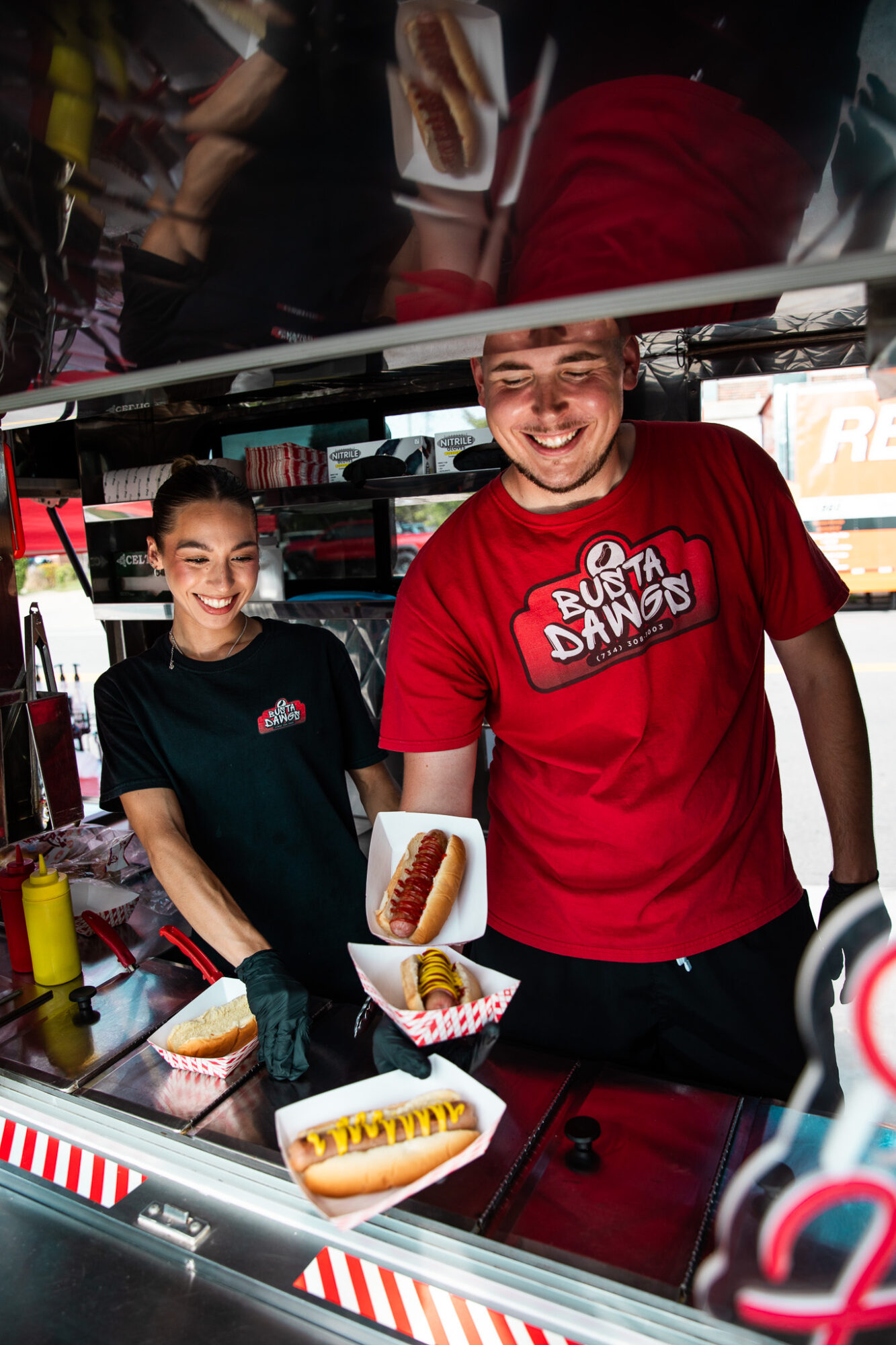 Two people behind a counter with food images, smiling, in a fast-food setting, one wearing a red shirt, the other in black.