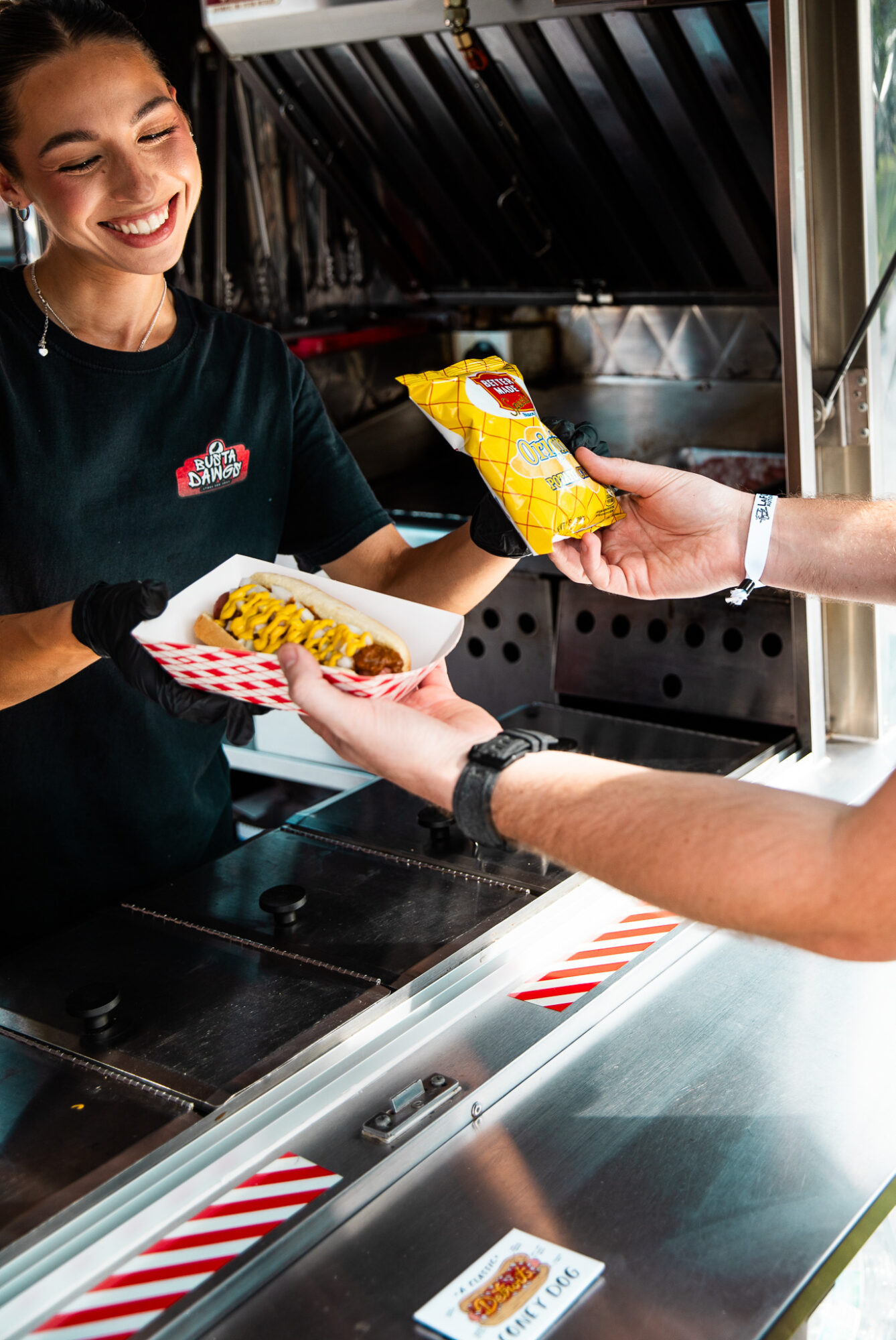 Smiling woman serving food at a counter, handing a plate to a customer holding a bag of chips.