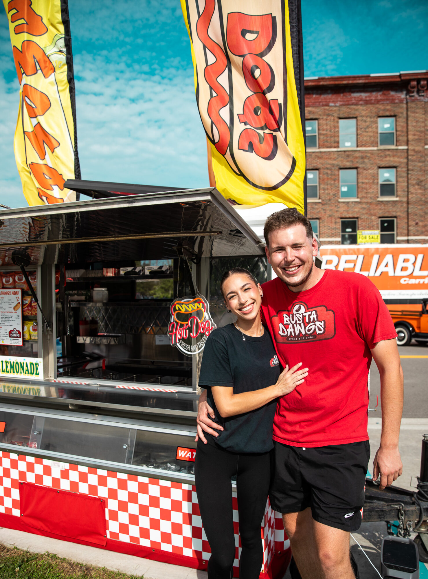A smiling young woman and man stand in front of a food truck with flags, in an urban setting.