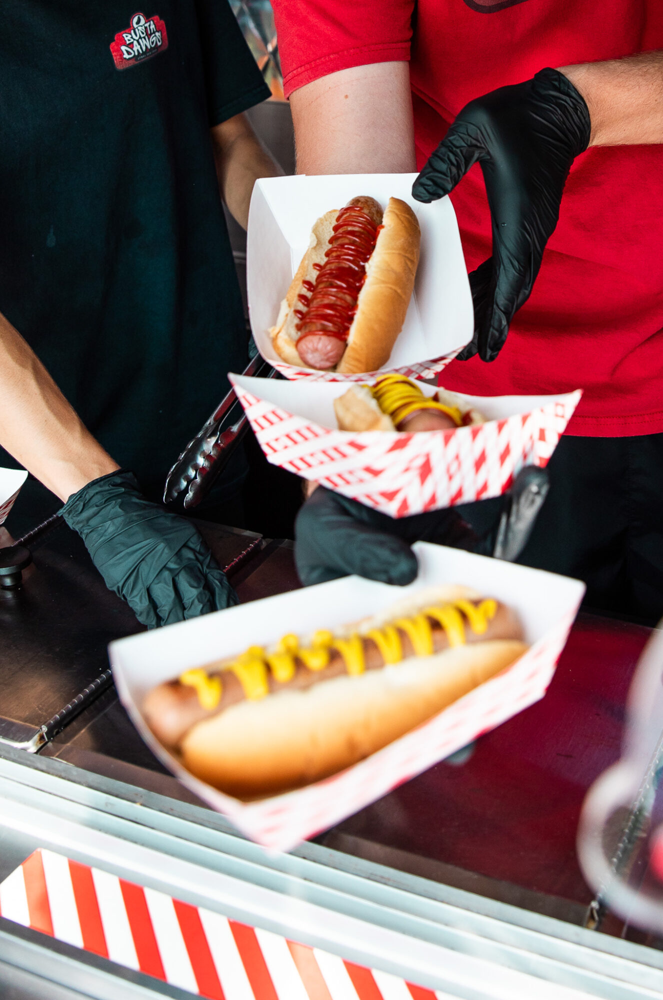Two people hold hot dogs in paper trays, one with ketchup and mustard, in a food stand. Word 'HOT DOG' is visible.