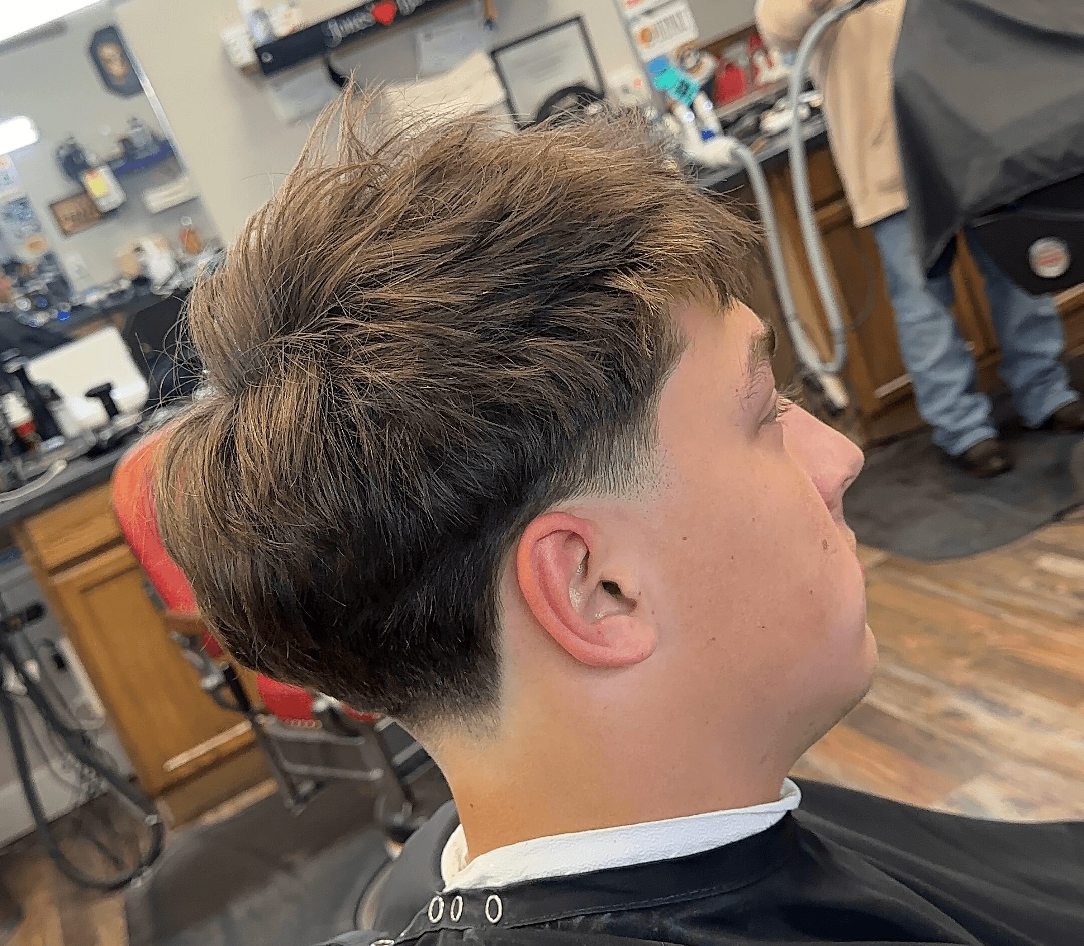 Young man with a fresh haircut in a barbershop, viewed from the side, with barber tools and chairs in the background.