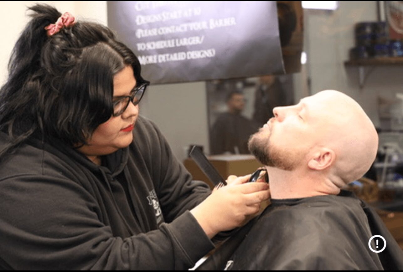 Woman with glasses and dark hair trims a man's beard in a barbershop, with a mirror and shelves in background.