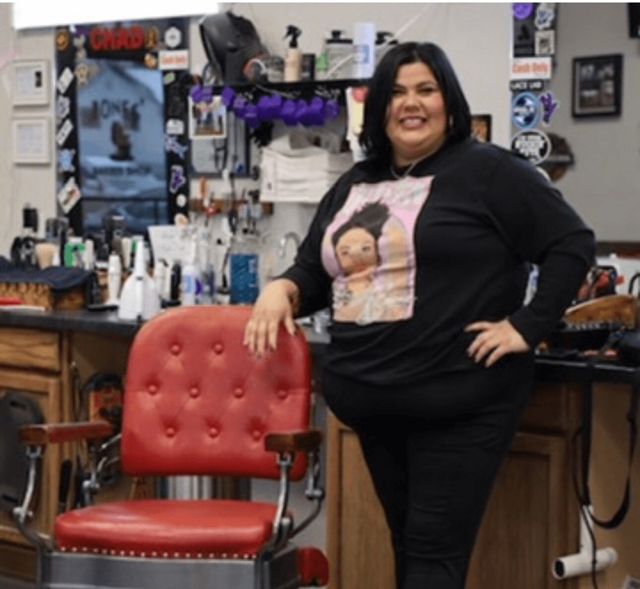 Woman with black hair smiling, standing in a barbershop, resting hand on red barber chair, surrounded by hair products and decorations.