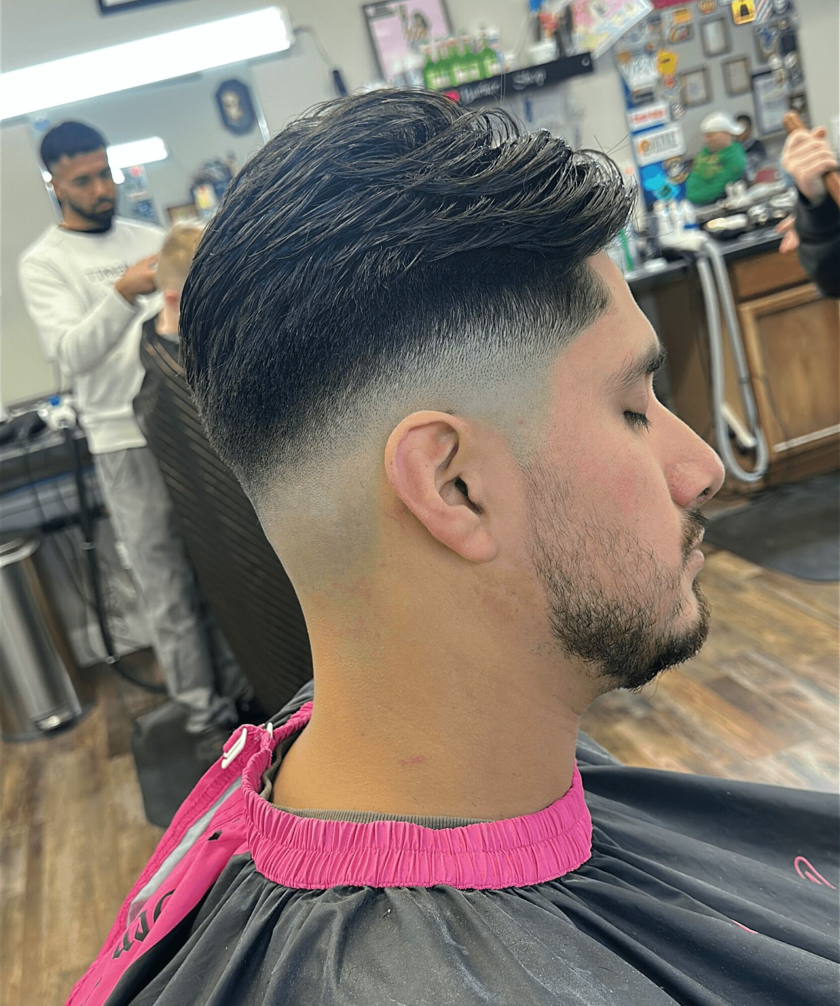 Young man with a fresh fade haircut, sitting in a barbershop, with a barber working in the background.