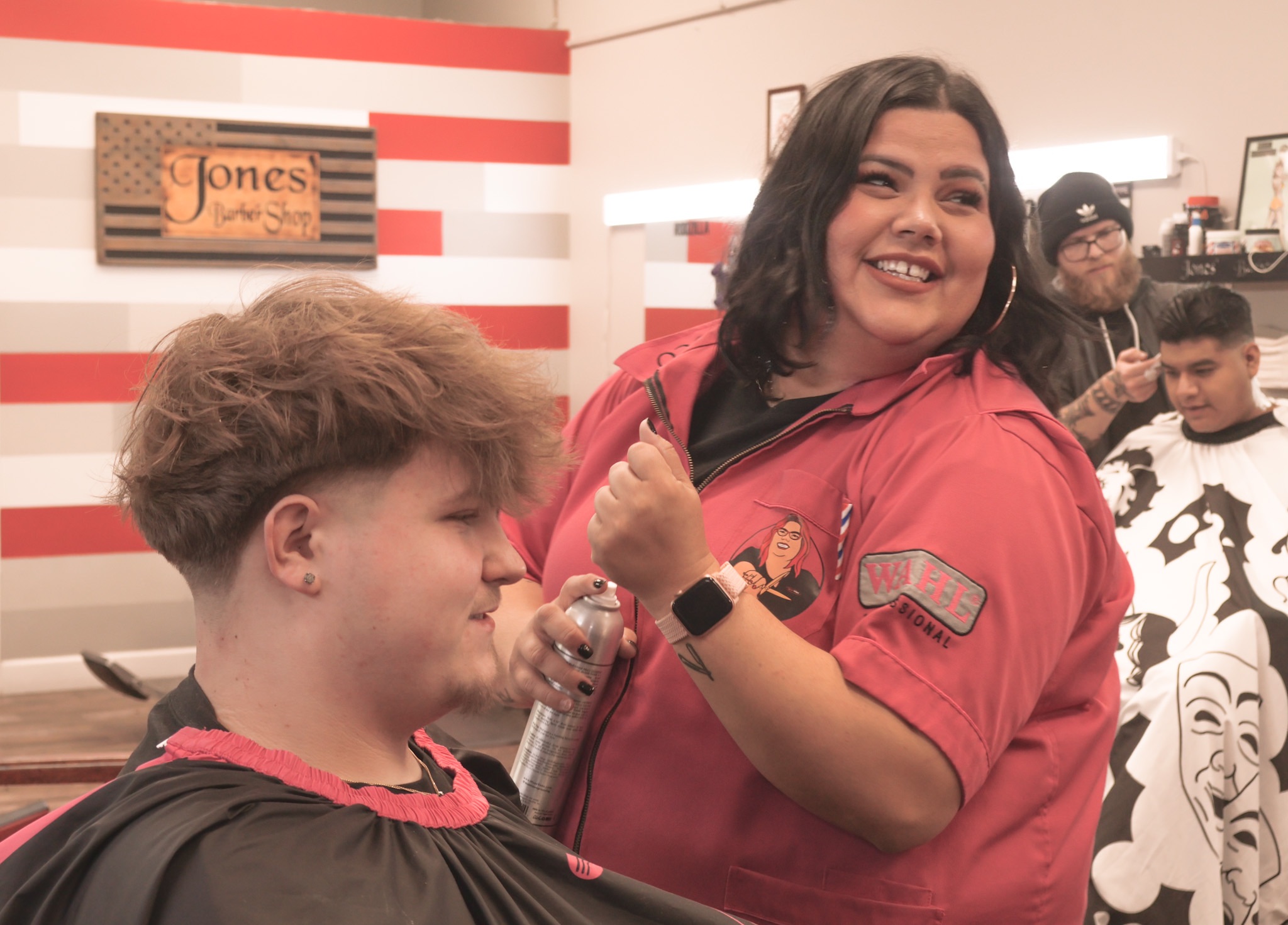 A woman with dark hair smiling and showing a fist bump to a young person with curly hair in a barber shop. Several people are in the background.