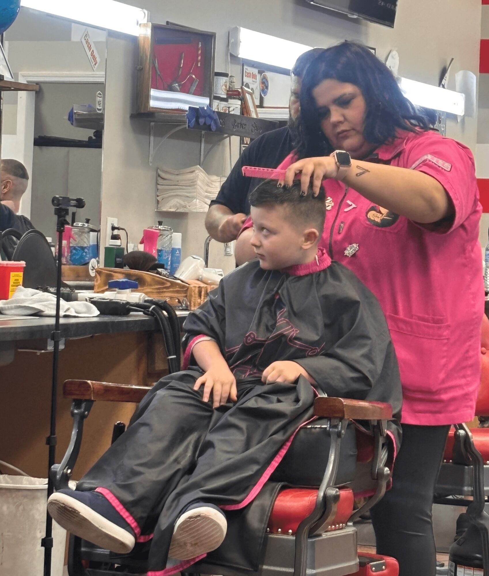 Barber cutting a child's hair in a salon, with hairdressing tools and mirrors visible in the background.
