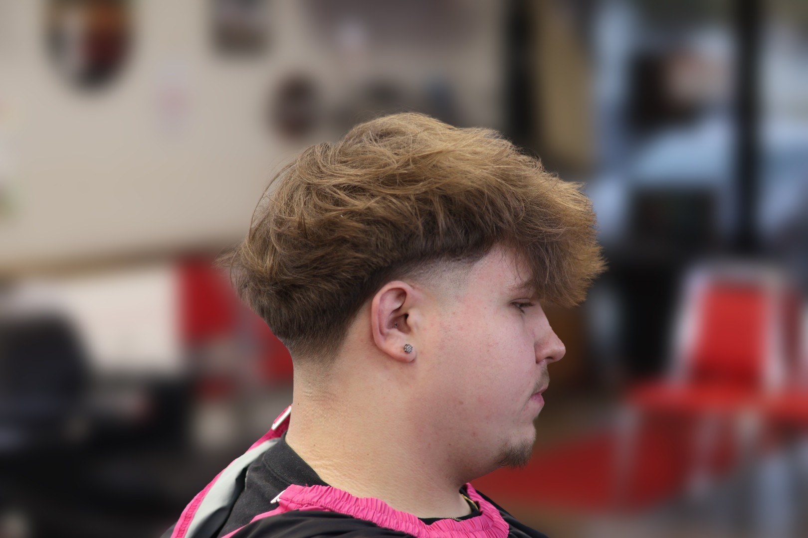 Young person with curly hair in a salon or barbershop, facing right, with blurred background of chairs and mirrors.
