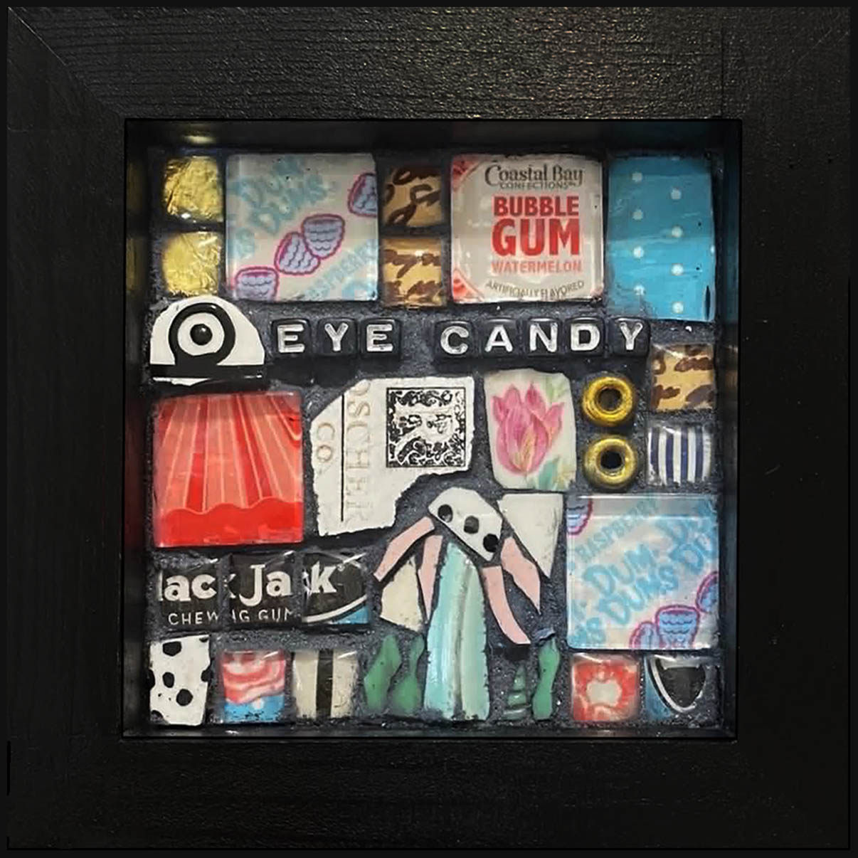 Display case with assorted candies and a paper cutout of a person, labeled 'Eye Candy', inside a black frame.