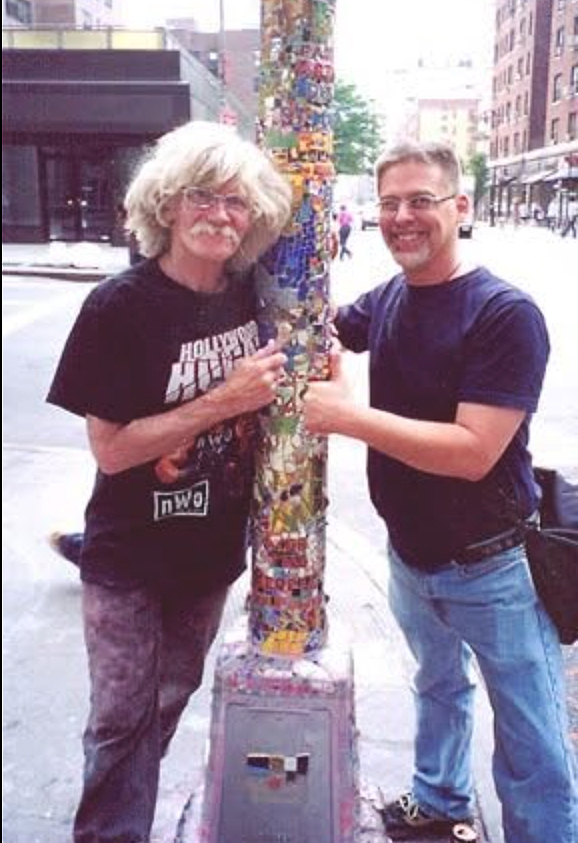 Two people stand outdoors next to a colorful pole covered in stickers, holding a small object together, smiling.