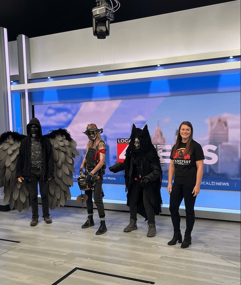 Four people in costumes stand on a stage with a blue screen background, one dressed as a wolf, others in dark costumes, and one in casual clothing.