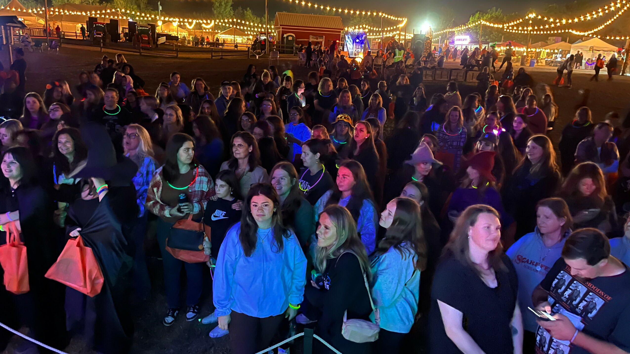 Crowd of people at an outdoor event with string lights and tents in the background, nighttime setting.