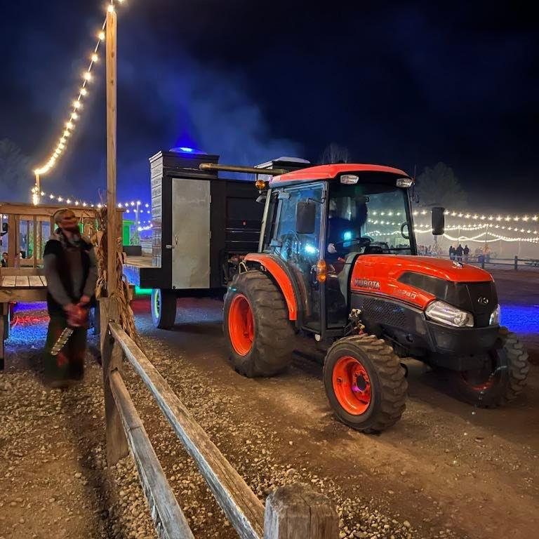 Red tractor with a trailer at night, illuminated string lights in the background, person standing nearby, dirt ground, dark sky.