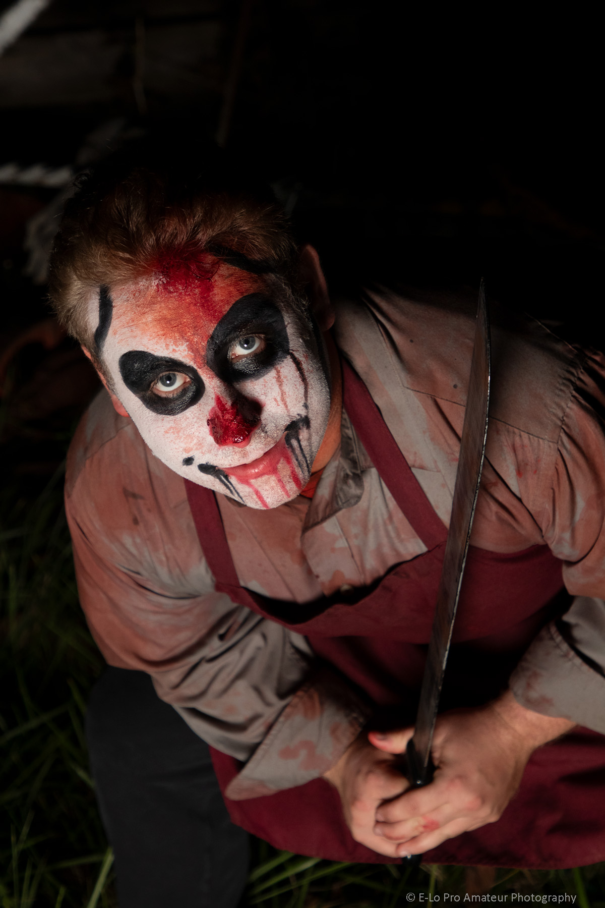 Person with face painted as a skull, holding a sword, wearing a beige shirt and maroon apron.