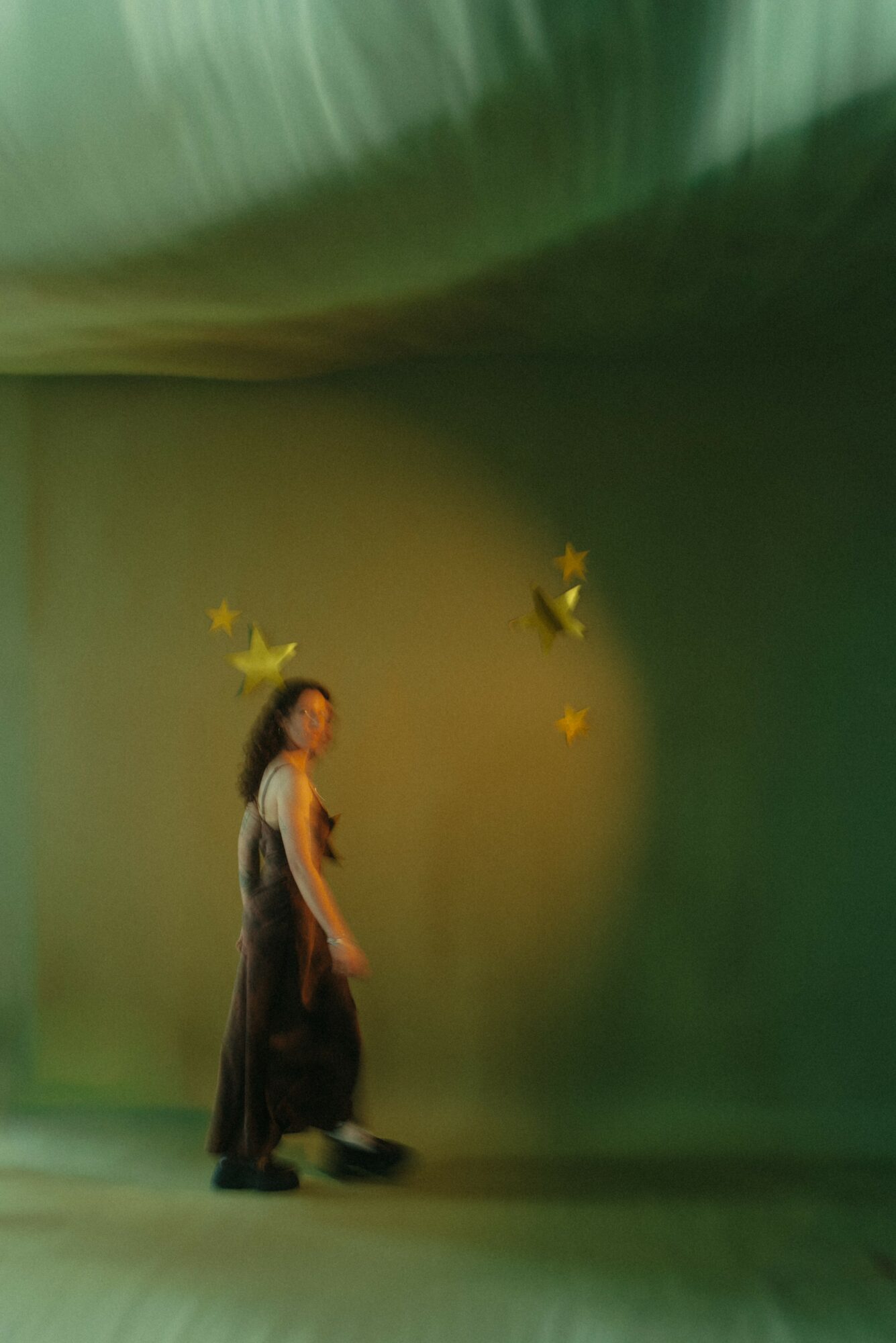A woman stands in front of a green background with yellow stars, looking to the side, with a blurred ceiling above.