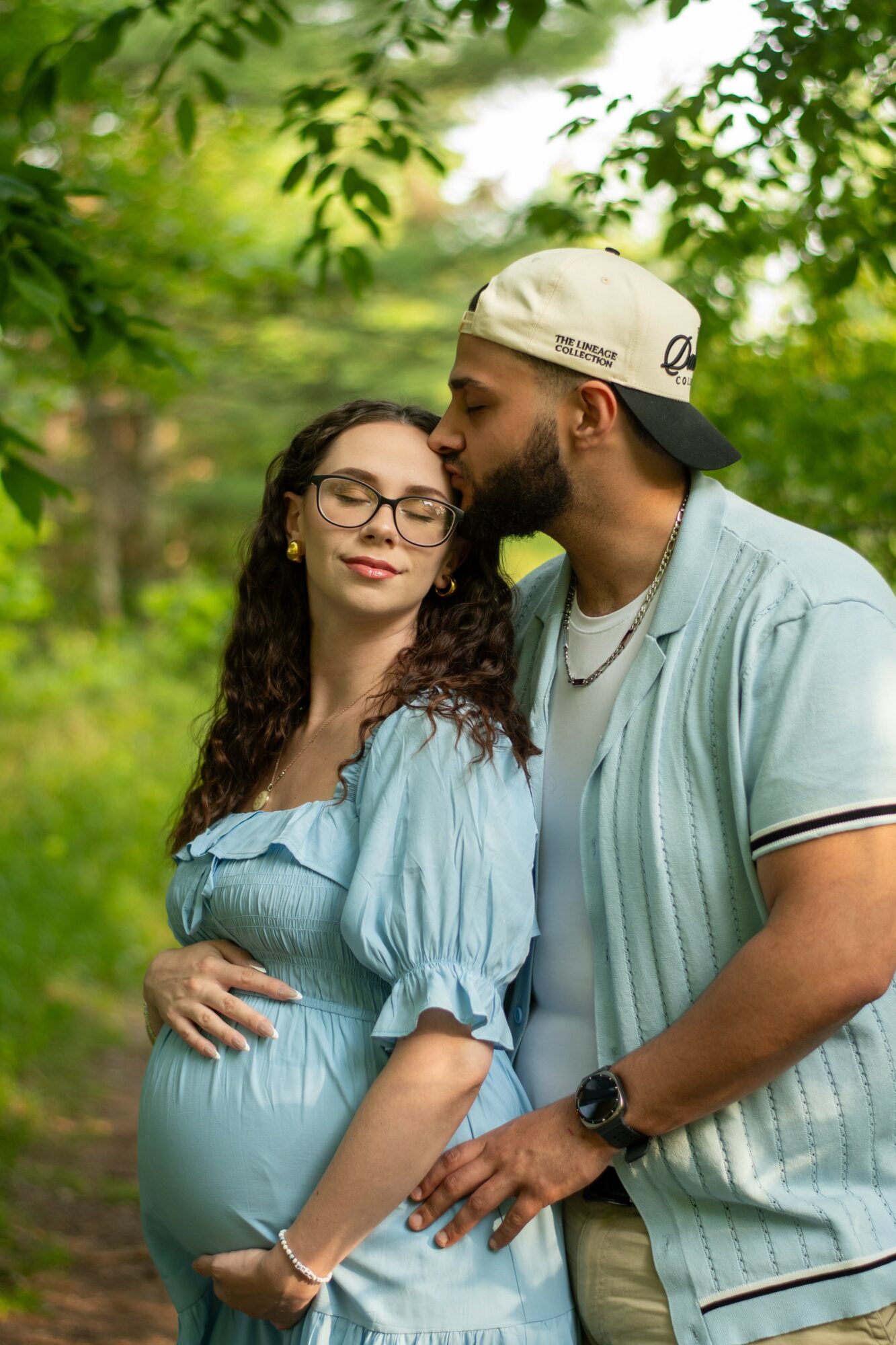 A pregnant woman with glasses and curly hair stands outdoors, while a man leans in to kiss her forehead, surrounded by green trees.