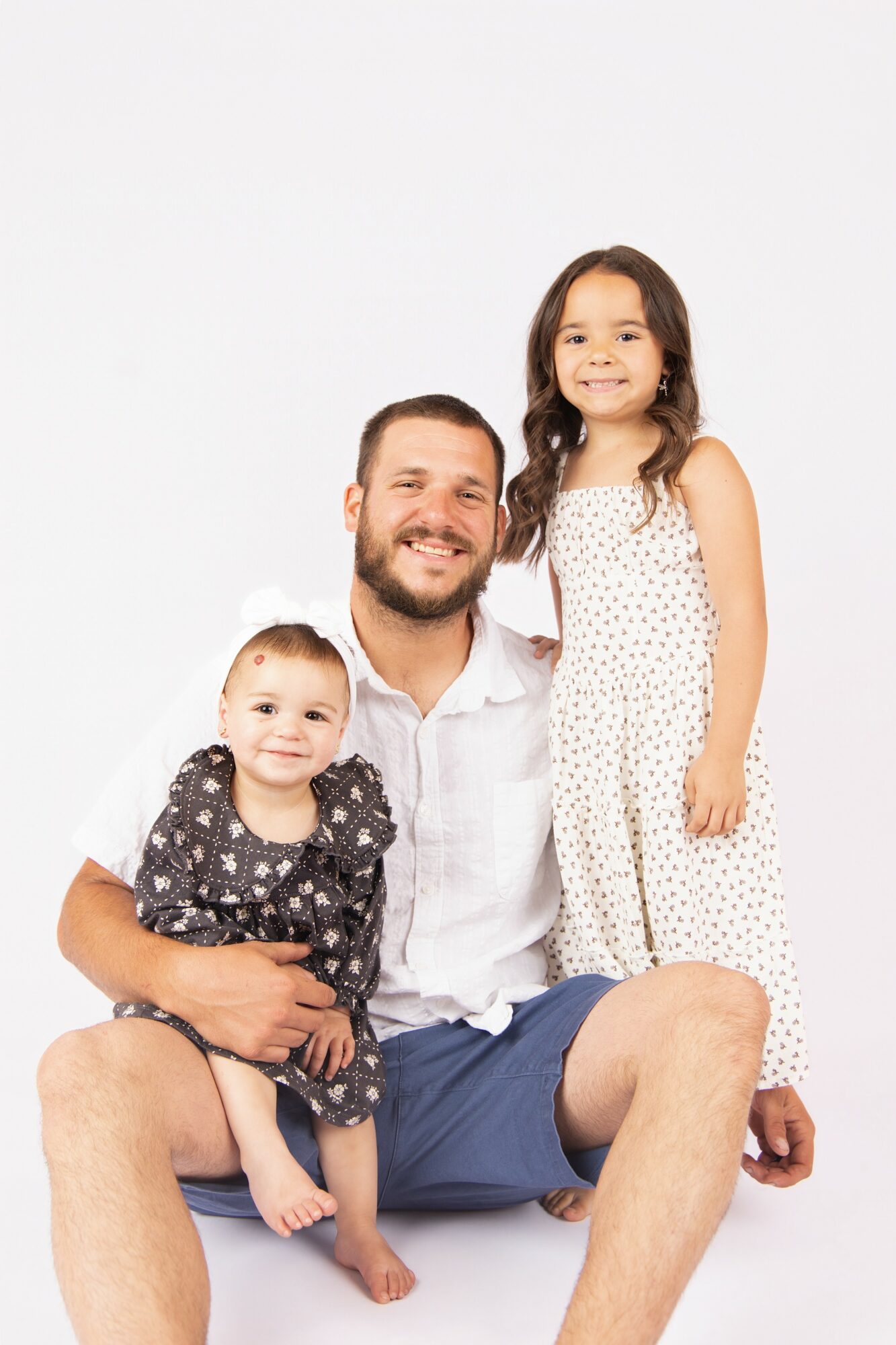 Man with two young girls, one sitting on his lap and the other standing beside him, smiling, against a plain white background.
