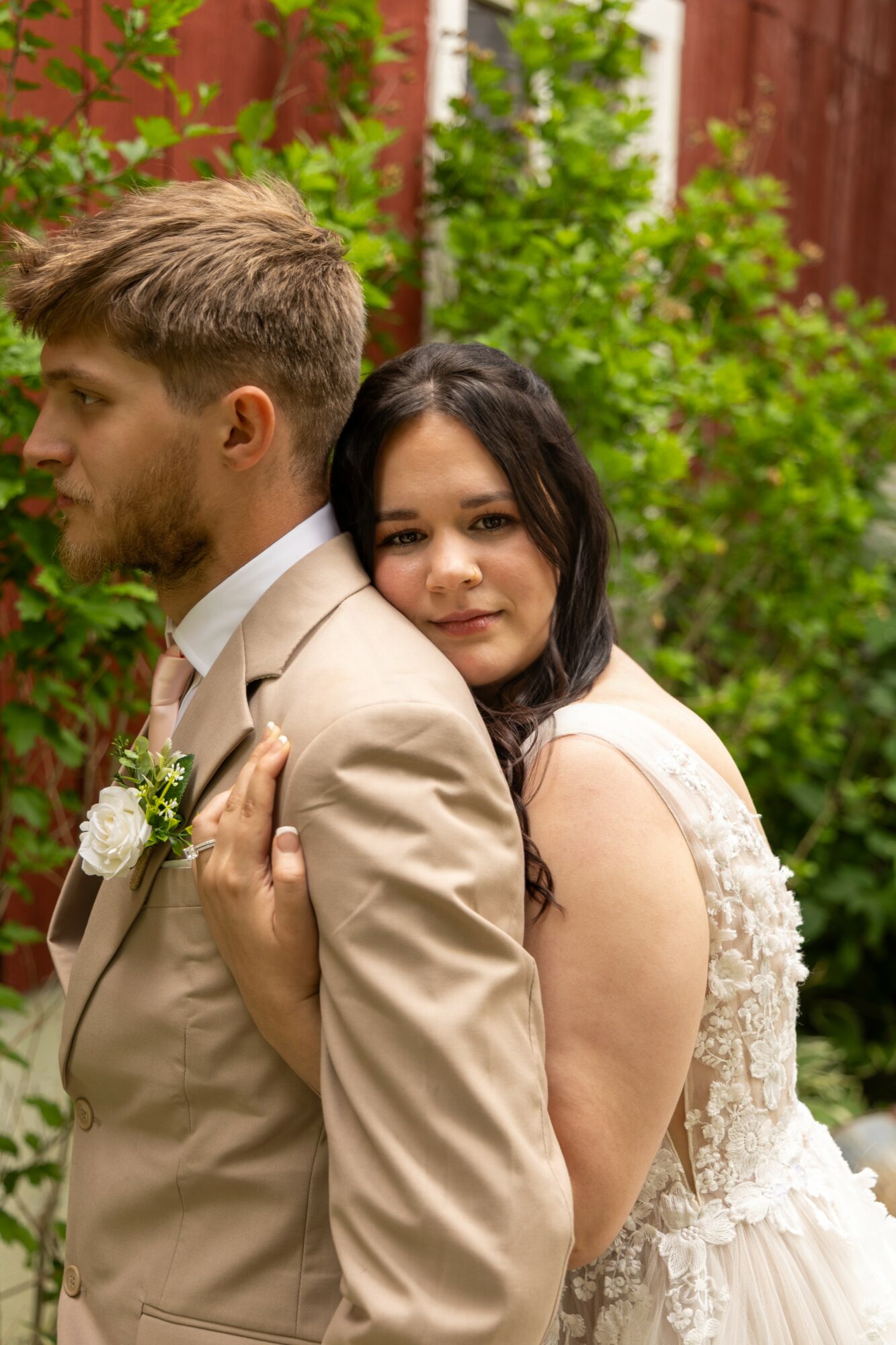 A woman in a wedding dress hugs a man in a beige suit outdoors, with green foliage and a red fence in background.