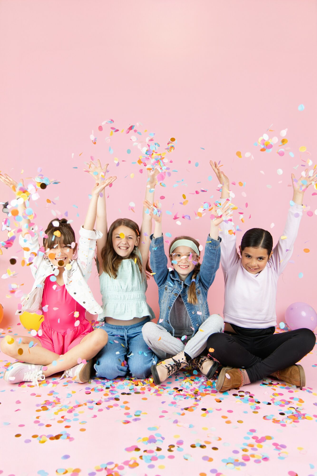 Four children sitting on a pink floor with a pink background, celebrating with confetti and balloons, smiling and raising hands.