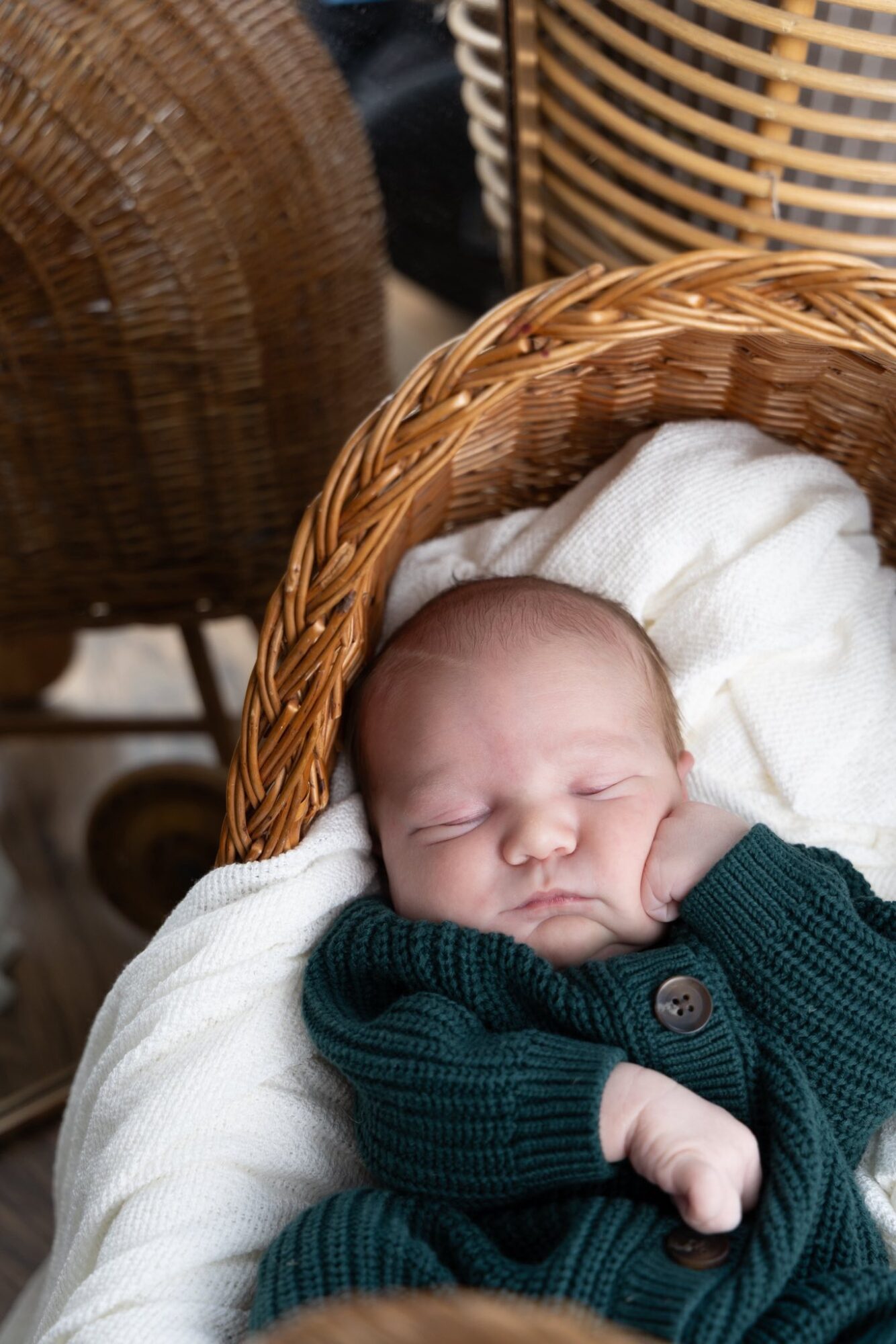 Baby sleeping in a wicker basket with a white blanket, wearing a dark green sweater, surrounded by woven baskets.