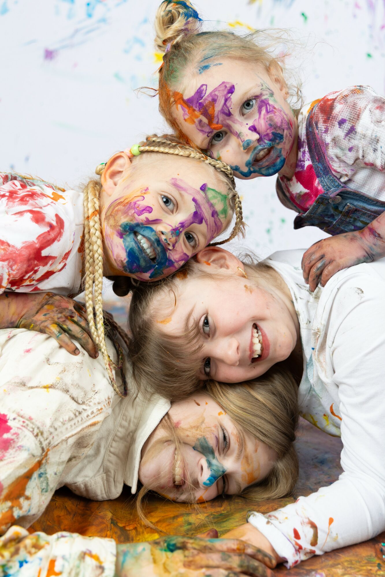 Four children with painted faces and colorful clothing lie together, smiling and playing on a white background.