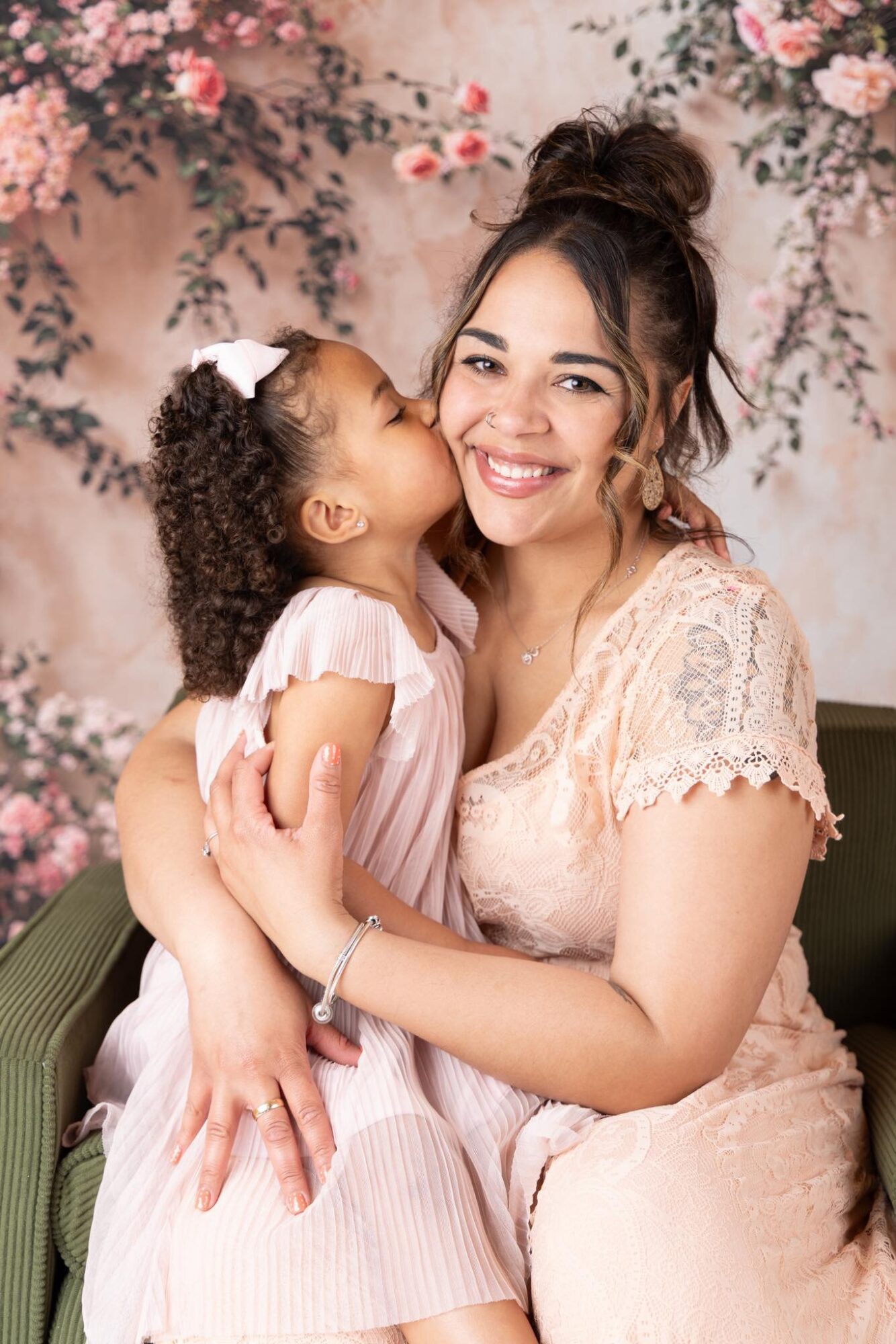 Young woman with dark hair in a bun smiling while holding a girl with curly hair and a white bow, against a floral background.