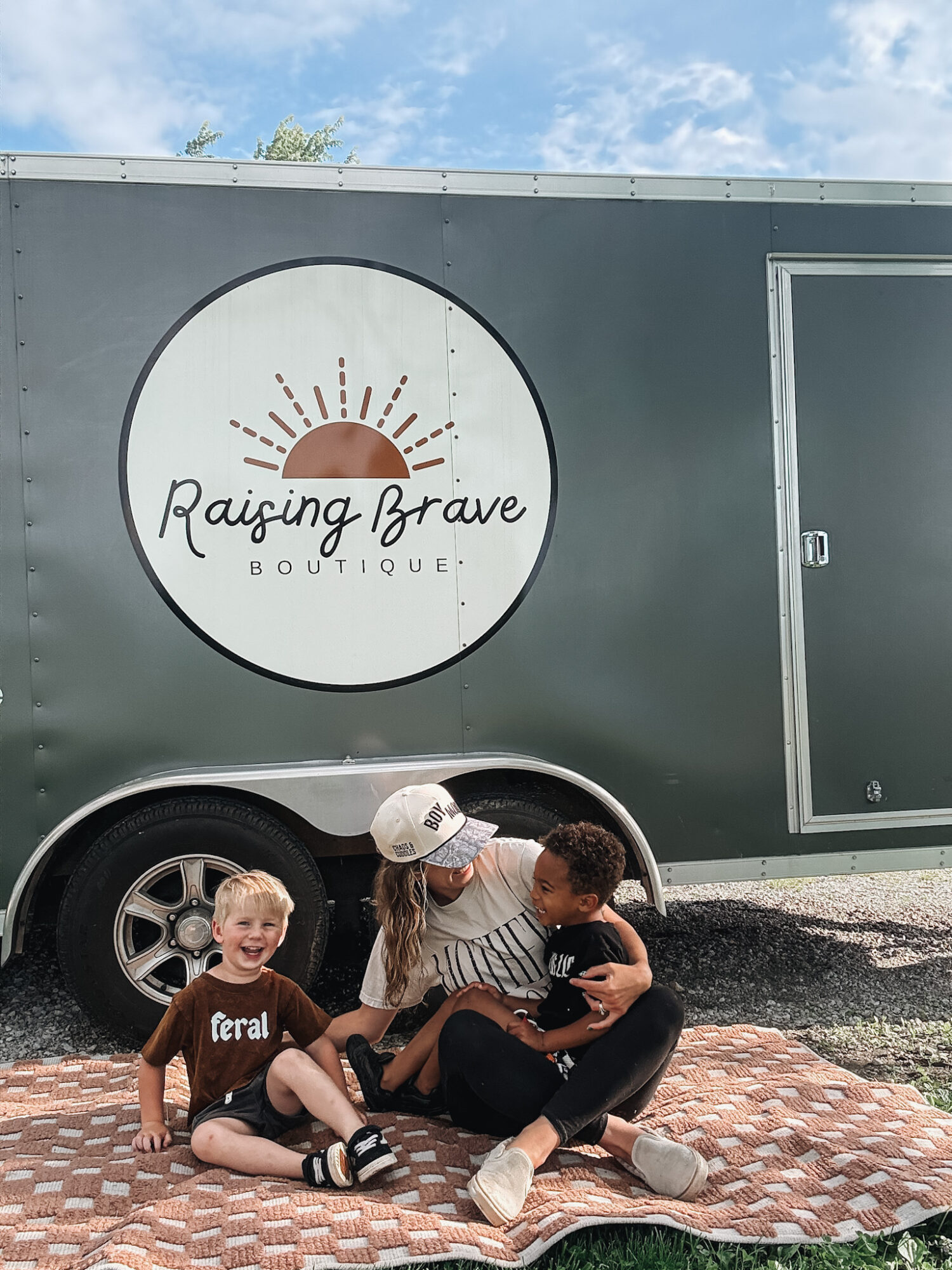 Three children sitting on a blanket outside, with a large circular sign on a vehicle behind them, under a blue sky.