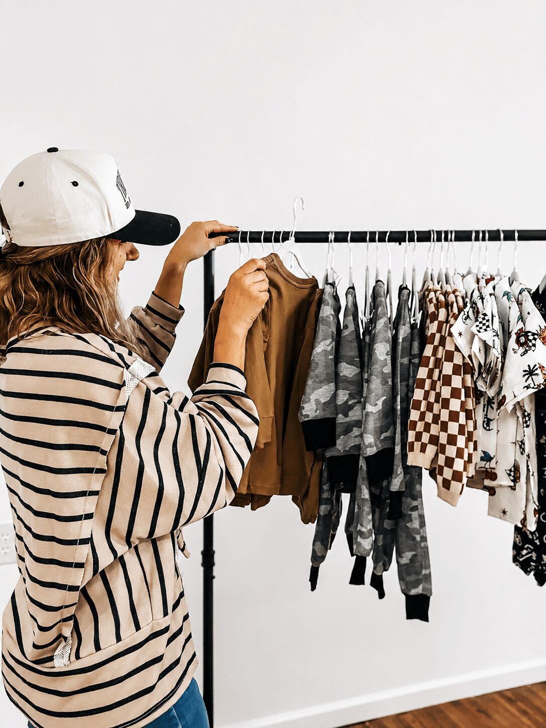 Person wearing a striped shirt and white cap looking at clothing on a rack, with a white wall background.