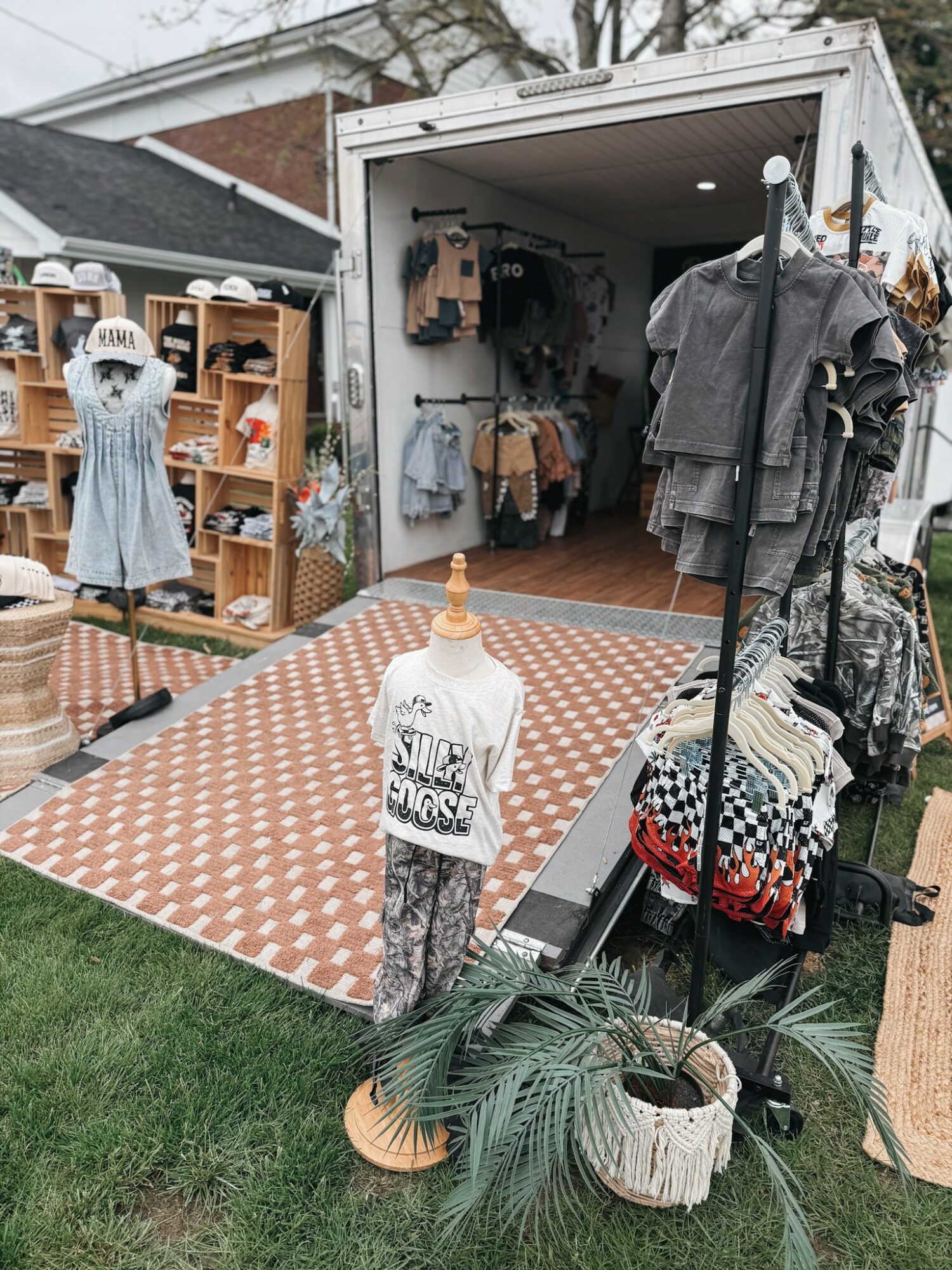 Outdoor clothing and accessories display with mannequins, racks, and shelves in front of a mobile shop, on a patterned mat.